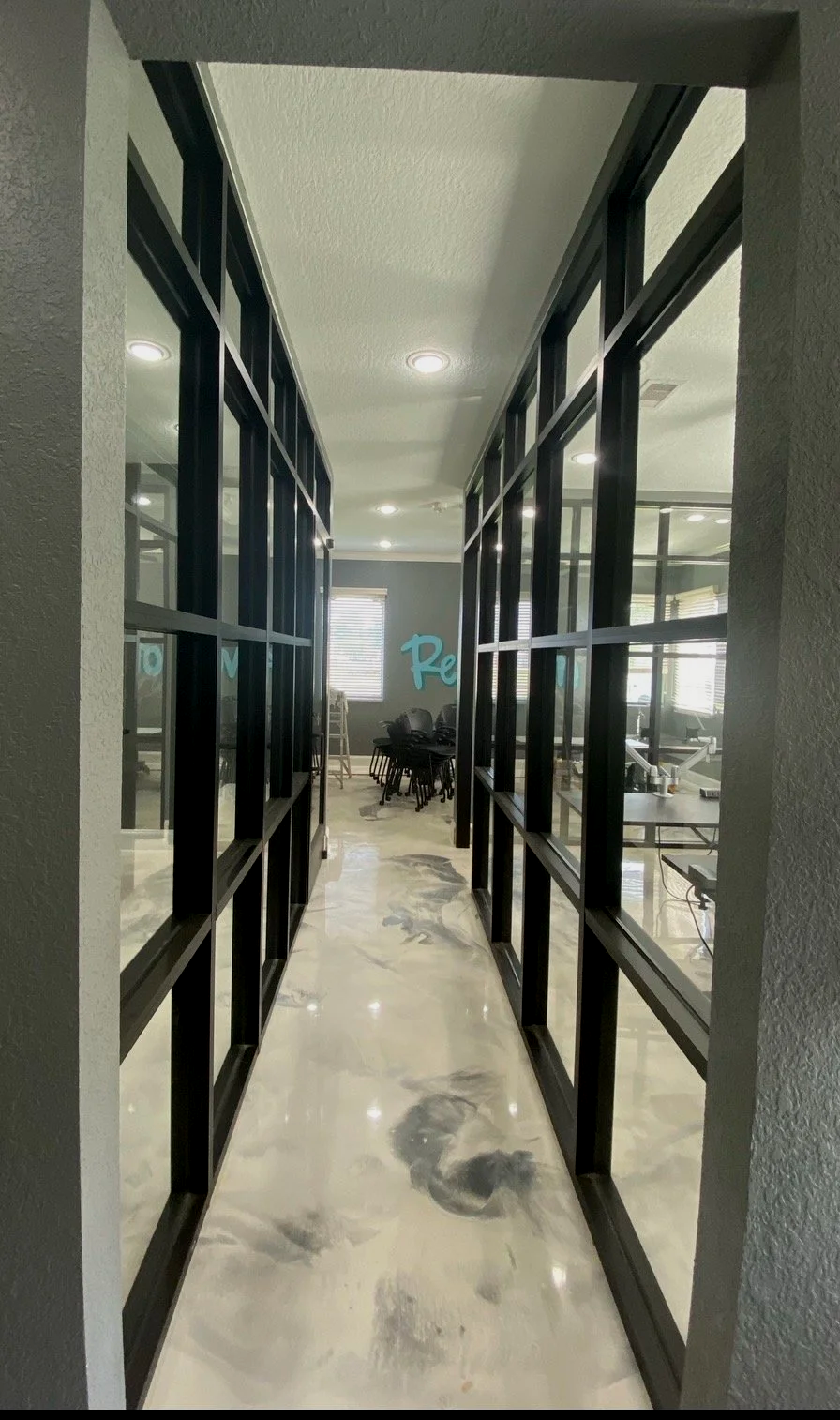 Photo of a hallway with black-framed glass partitions on both sides and a marble floor, leading to a room with chairs and windows in the background.