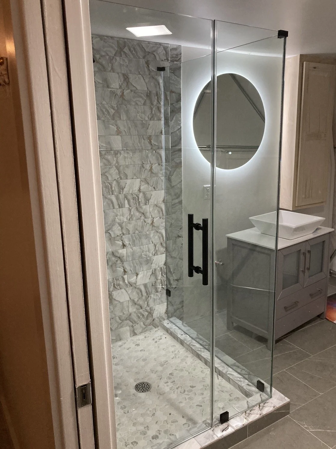 Modern glass-enclosed shower with gray marble tile walls and pebble tile floor, adjacent to a bathroom vanity with a vessel sink and a circular illuminated mirror.