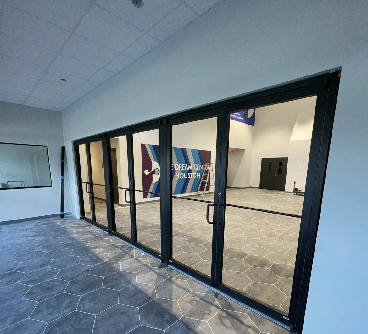 Interior view of the Dream Center Houston, showing glass doors leading to an empty room with a ladder, a colorful mural, and a sign reading "Dream Center Houston."