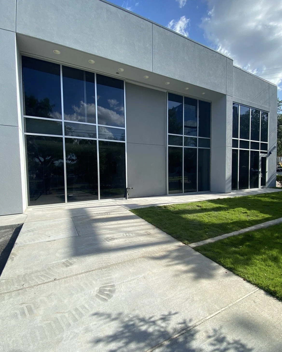 Modern building with large reflective glass windows, a concrete walkway, and a small grassy area in front, under a partly cloudy sky.