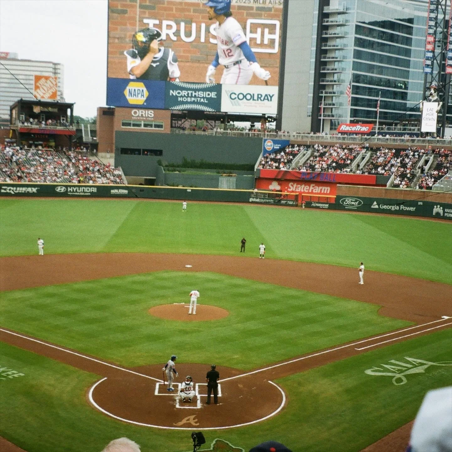 Film photo of a baseball stadium from the stands, showing the field with players positioned on the bases, the batter at home plate, and the large digital scoreboard with advertisements and an image of two baseball players.