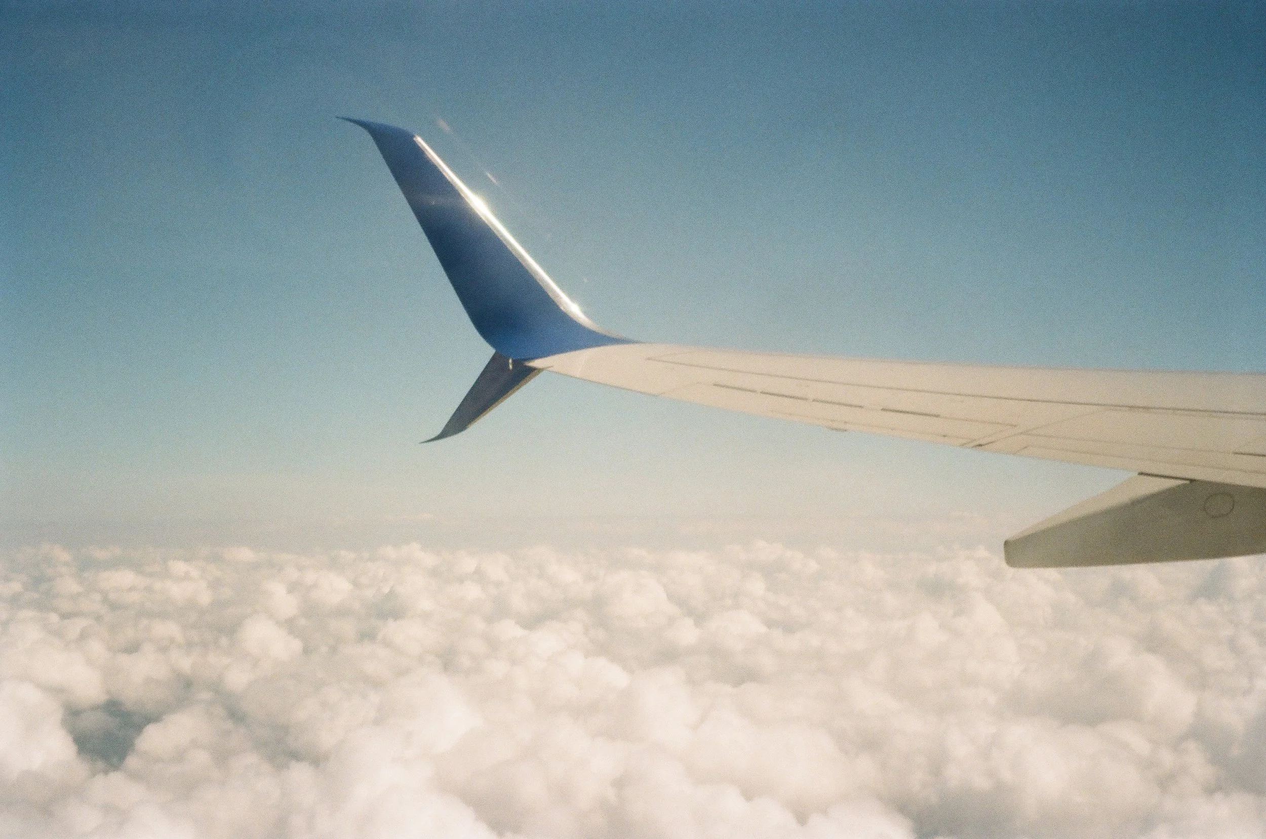 View from an airplane window showing the wing with a blue wingtip flying above white clouds under a clear blue sky.