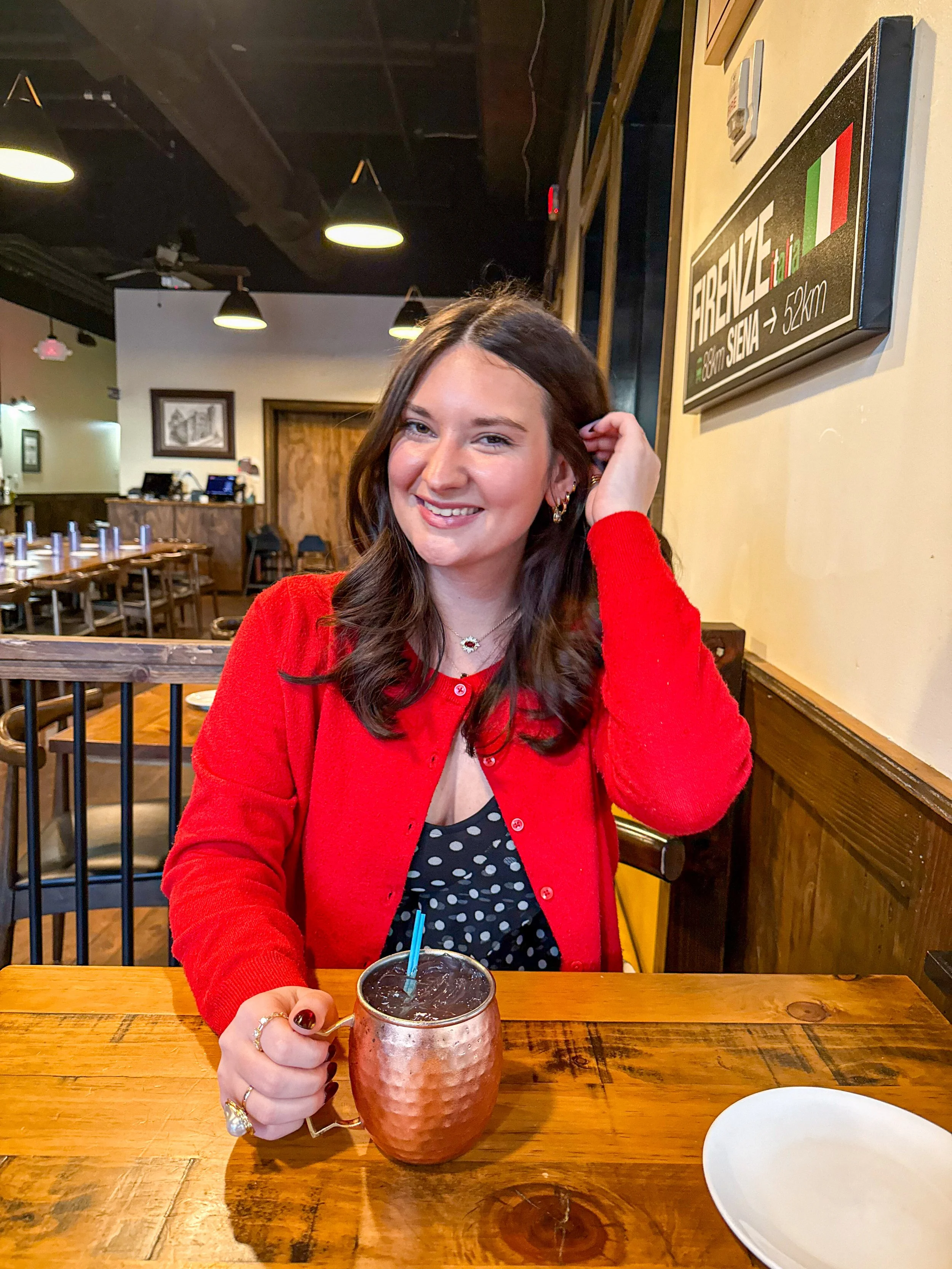 A photo of Marlee, wearing a red cardigan and a black dress with white polka dots, sitting at a wooden table in a restaurant, smiling at the camera.