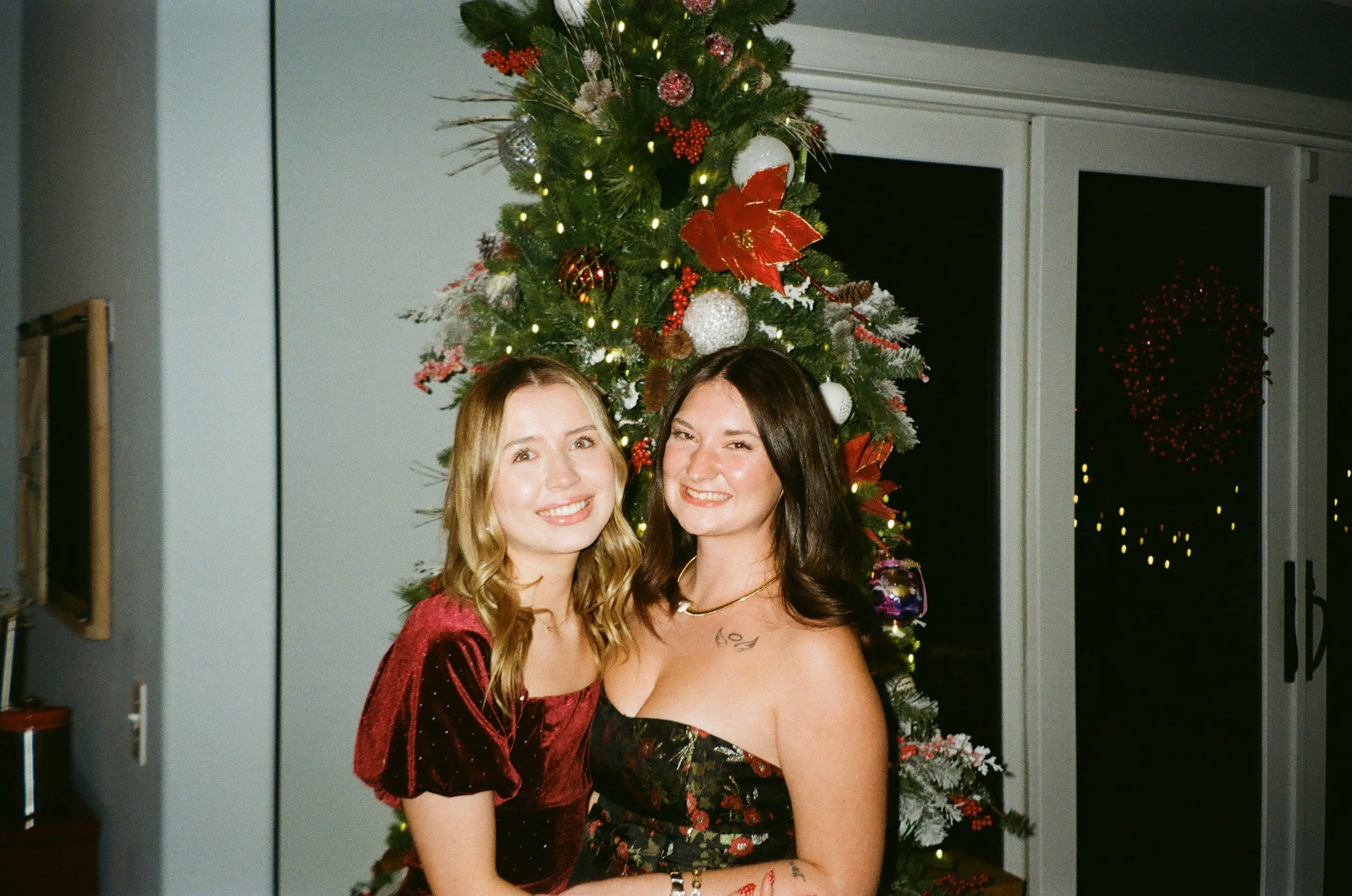 A film photo of Marlee and a friend smiling in front of a decorated Christmas tree at night.