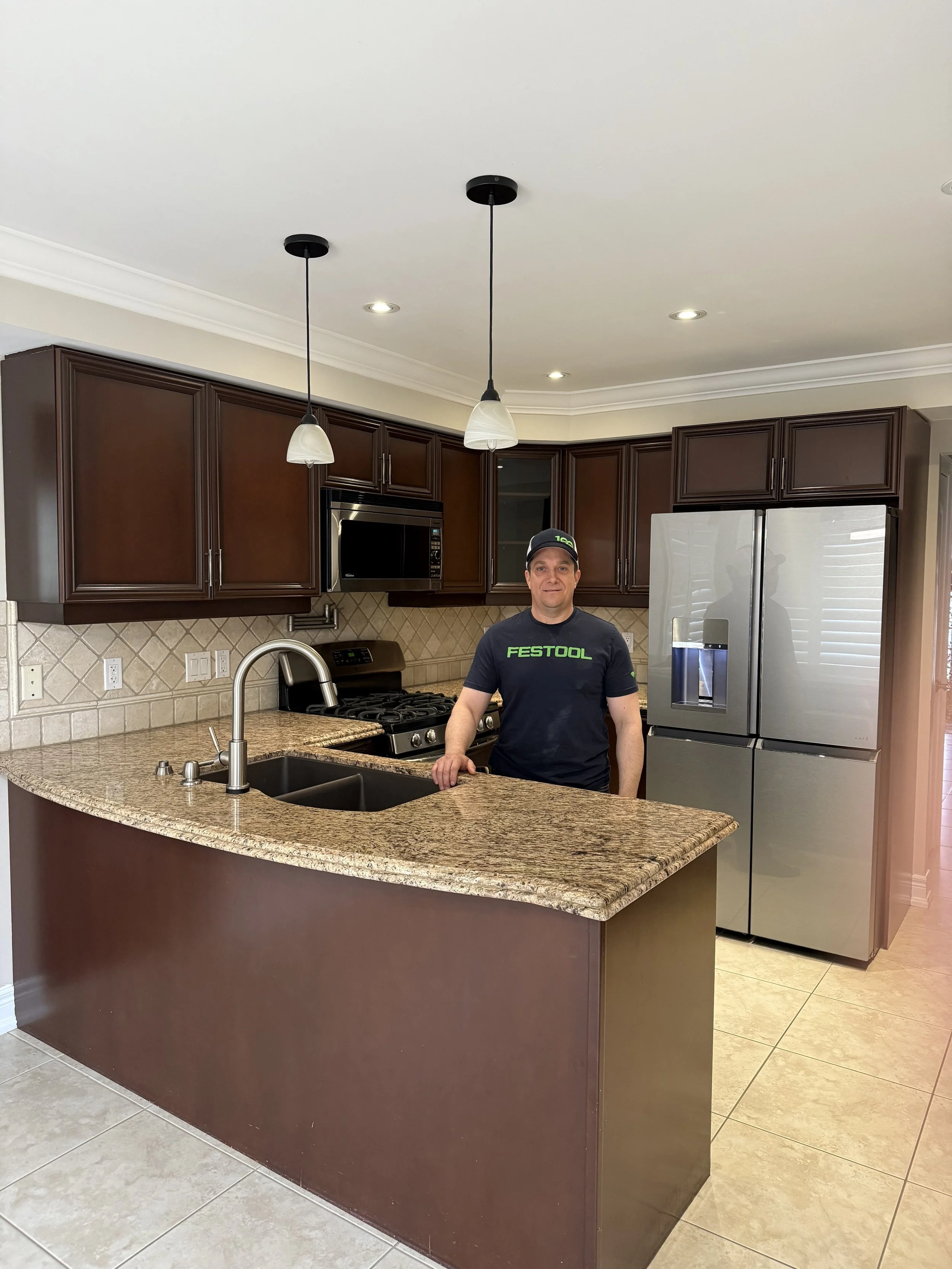 A man standing in a kitchen with wooden cabinets, granite countertops, a stainless steel refrigerator, and a black microwave. He is wearing a black Festool T-shirt and cap, and there is a double sink and stovetop visible.