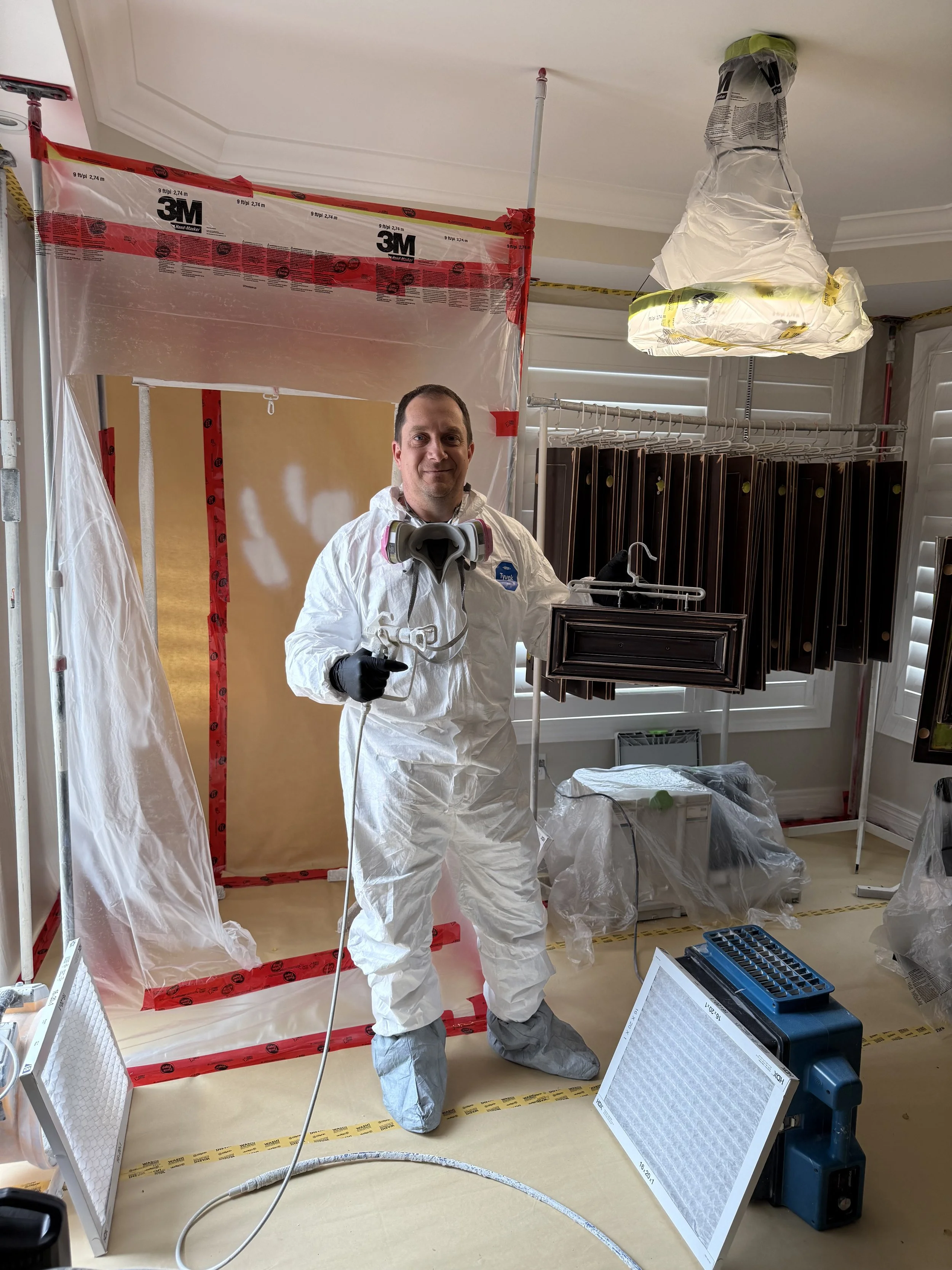 A man in a white protective suit holding a spray gun stands in a construction or renovation site with plastic sheeting and protective materials around.