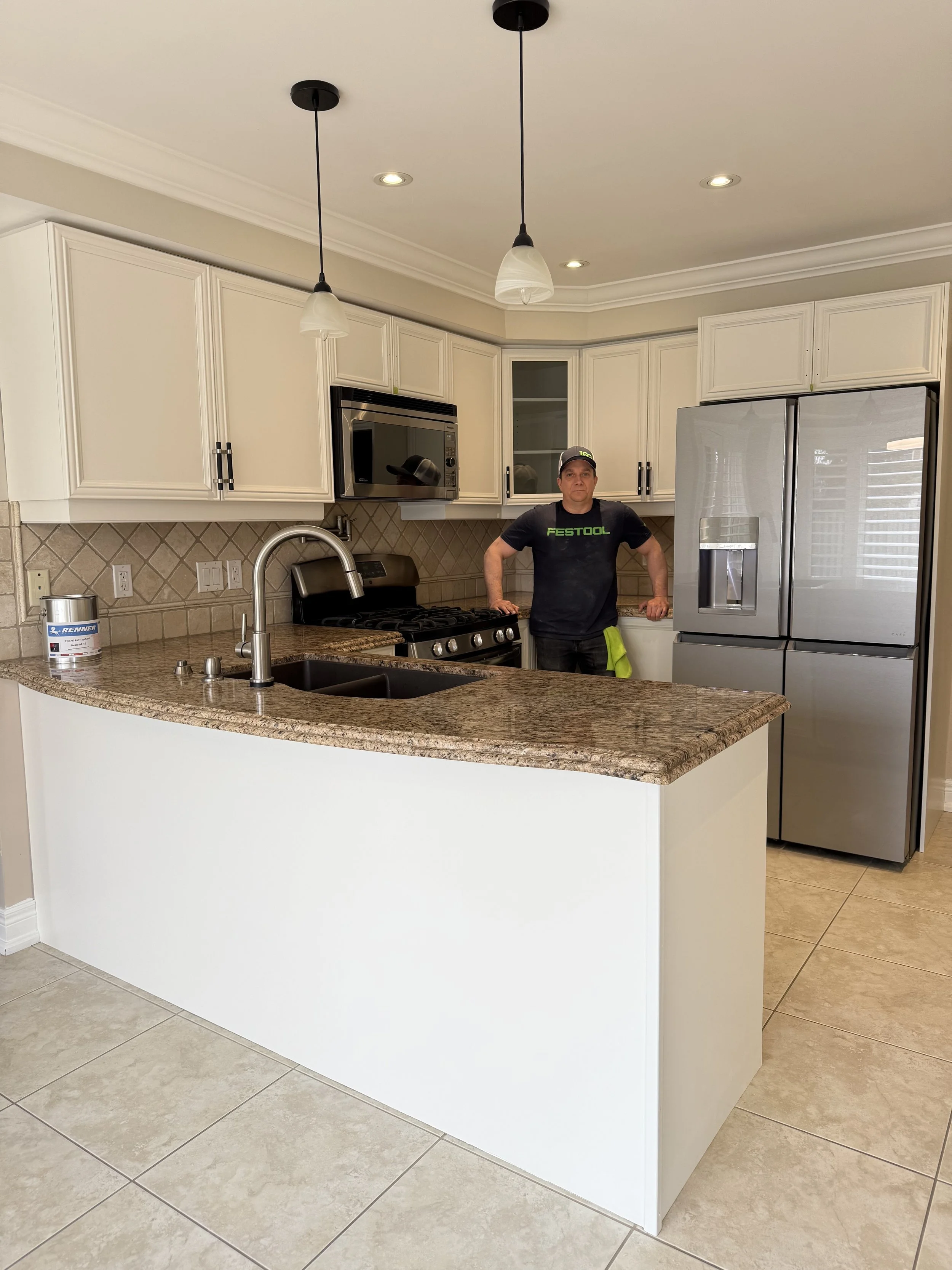 A man wearing a black Festool t-shirt stands behind a kitchen island with a granite countertop in a modern kitchen. The kitchen features white cabinets, a stainless steel refrigerator, stove, microwave, and tiled backsplash. The ceiling has recessed 