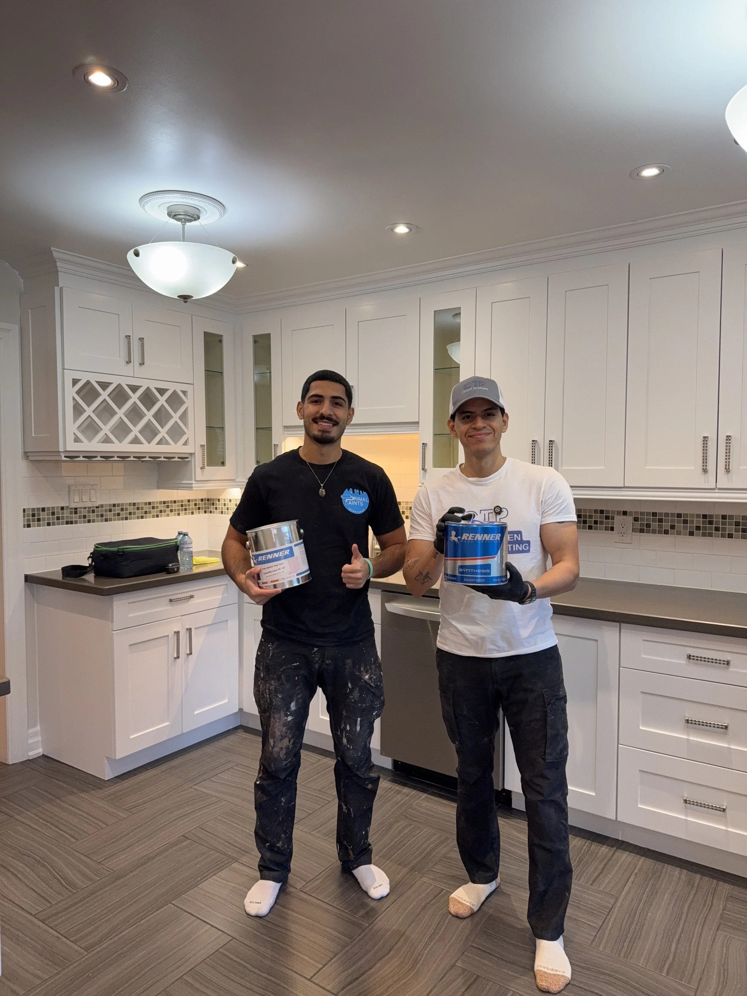 Two men standing in a kitchen holding paint cans and smiling. They are wearing work clothes with paint stains and socks.