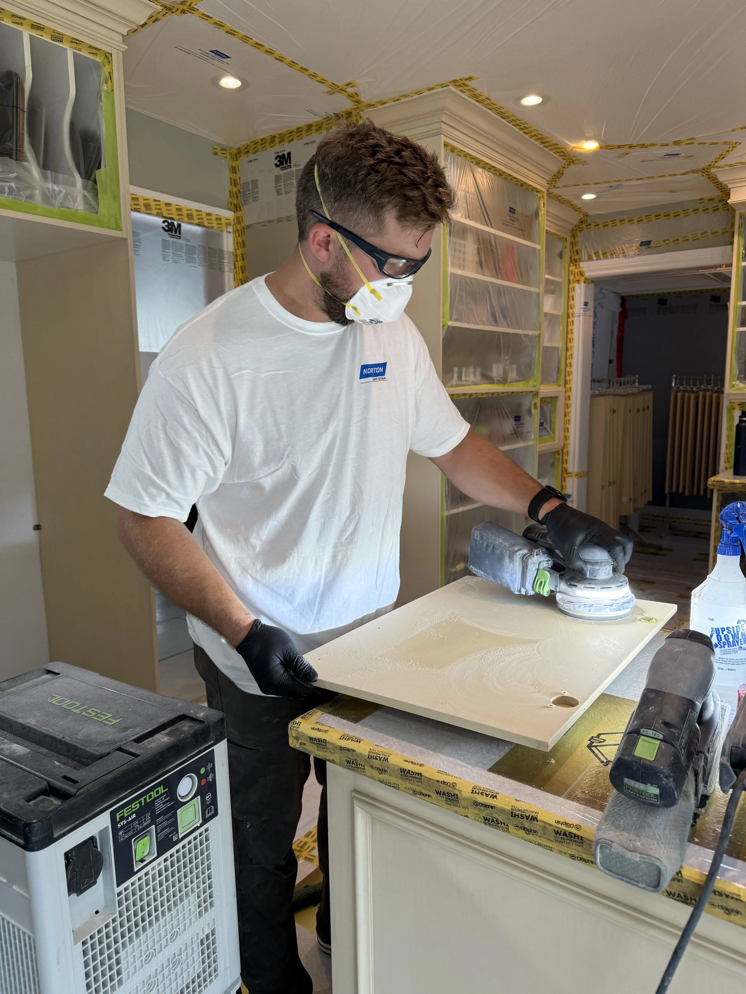 A man wearing a white t-shirt, safety glasses, and a face mask is sanding a piece of wood using an electric sander in a room under construction.