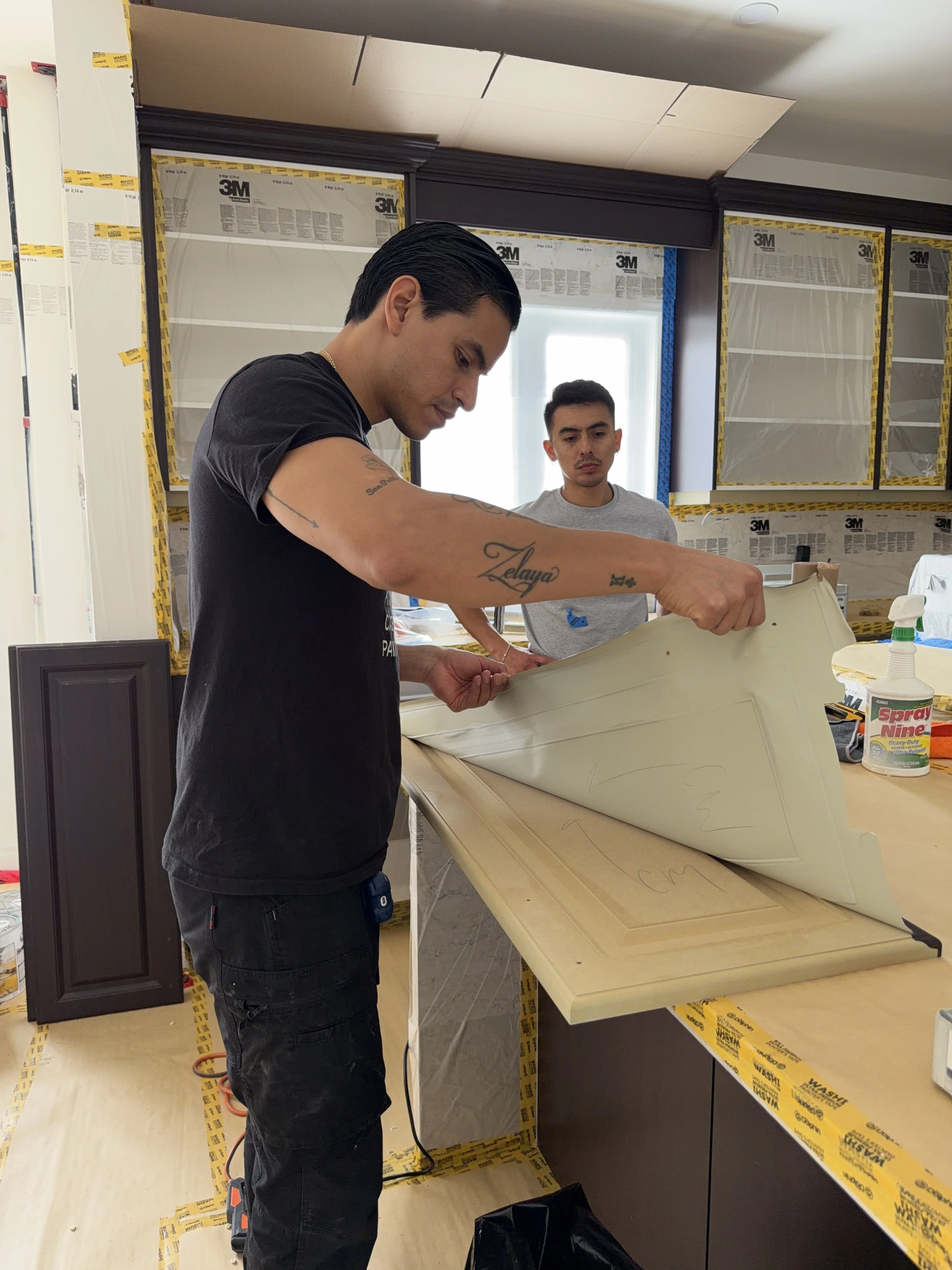 Two men working on a woodworking project in a workshop, with one lifting or pulling back a large piece of foam or insulation, and the other observing in the background.
