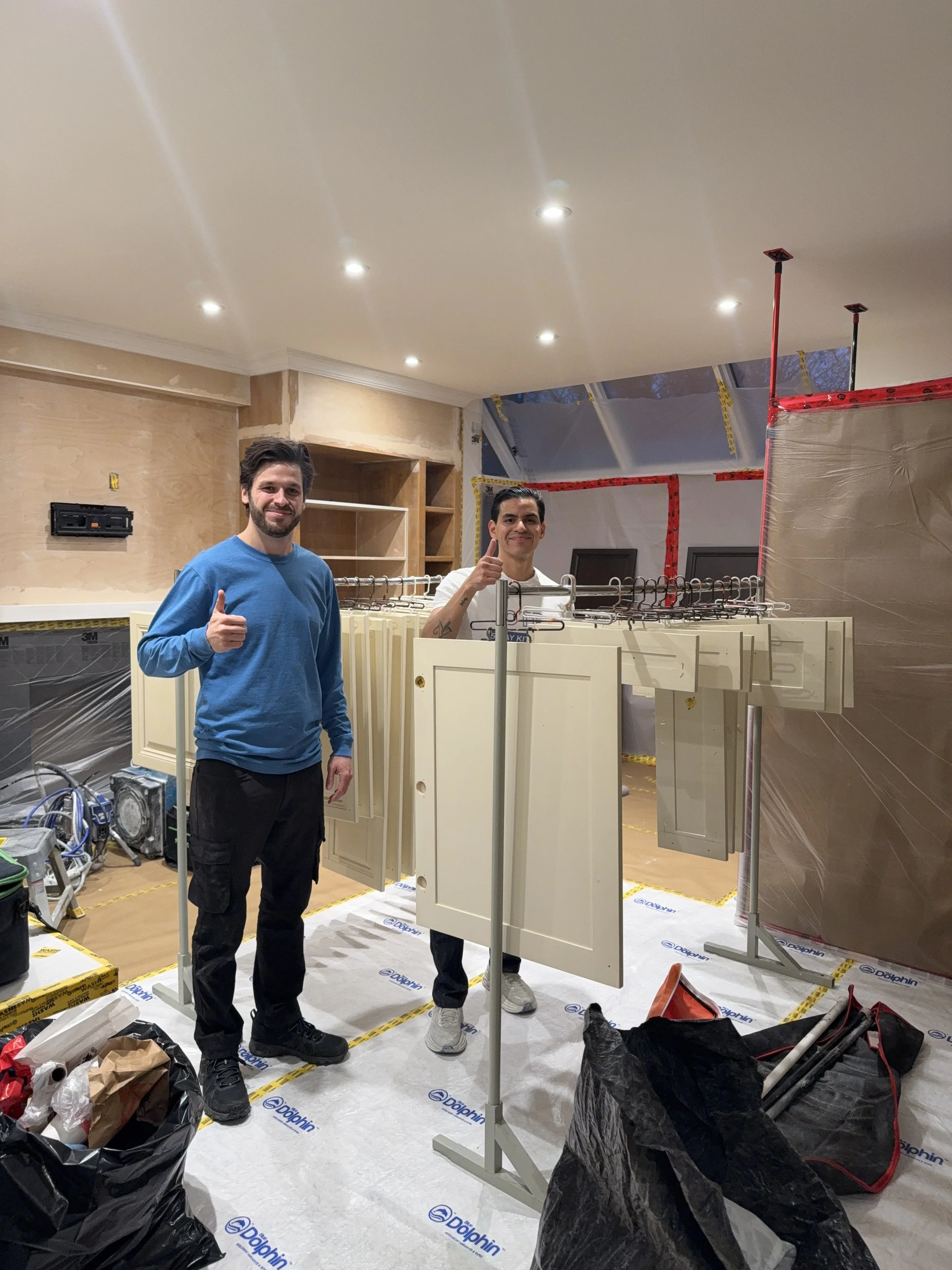 Two men standing in a room under construction, giving thumbs up, with kitchen cabinets and construction materials around.