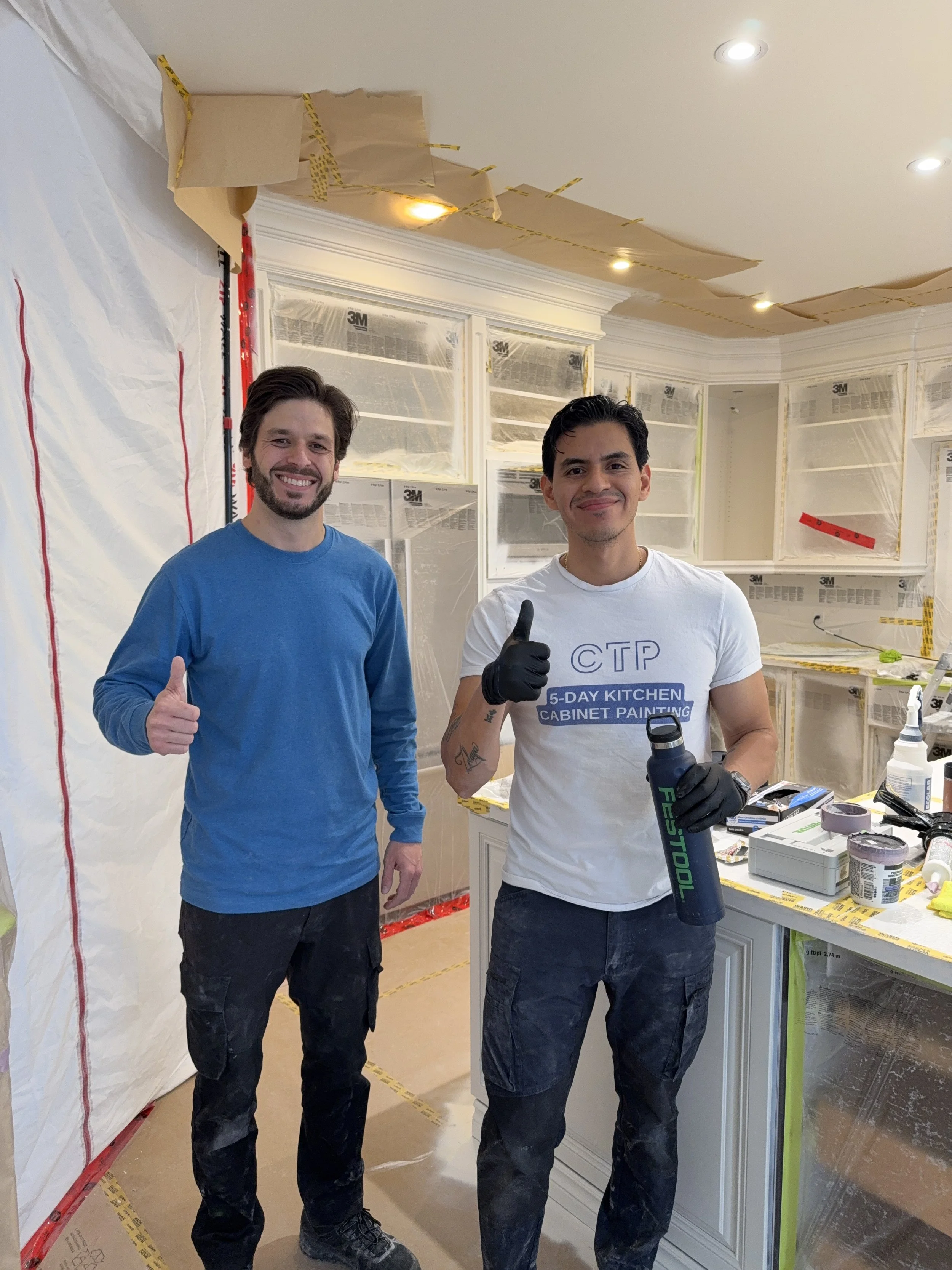 Two men standing in a kitchen under renovation, giving thumbs-up. They are wearing work clothes and gloves, surrounded by painting supplies and plastic-covered cabinets, with repair tape and paper on the walls and ceiling.