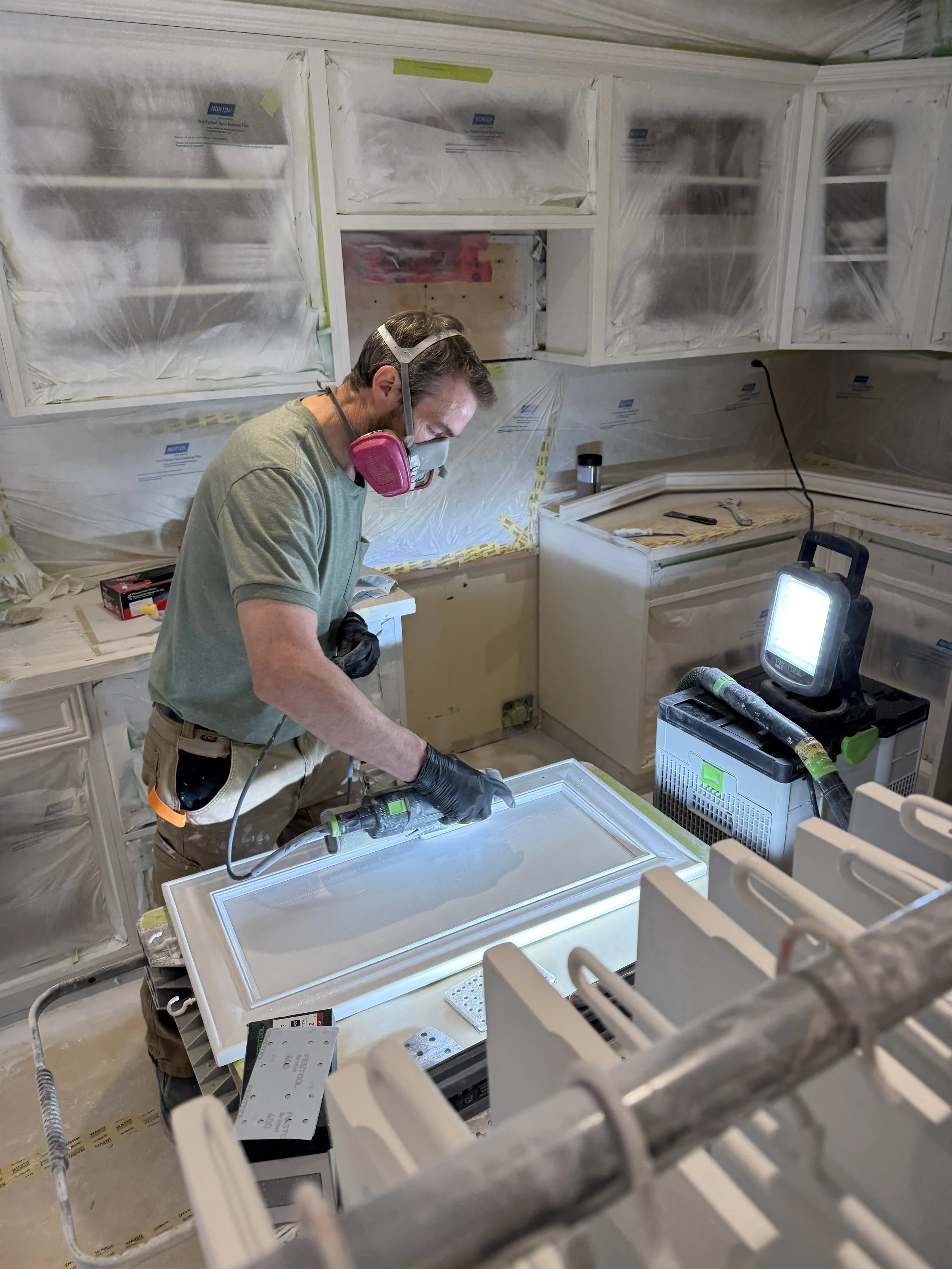 A man wearing a pink respirator mask and black gloves working on a cabinet door using a tool, in a kitchen under renovation with plastic covering on the cabinets and yellow tape marking areas.
