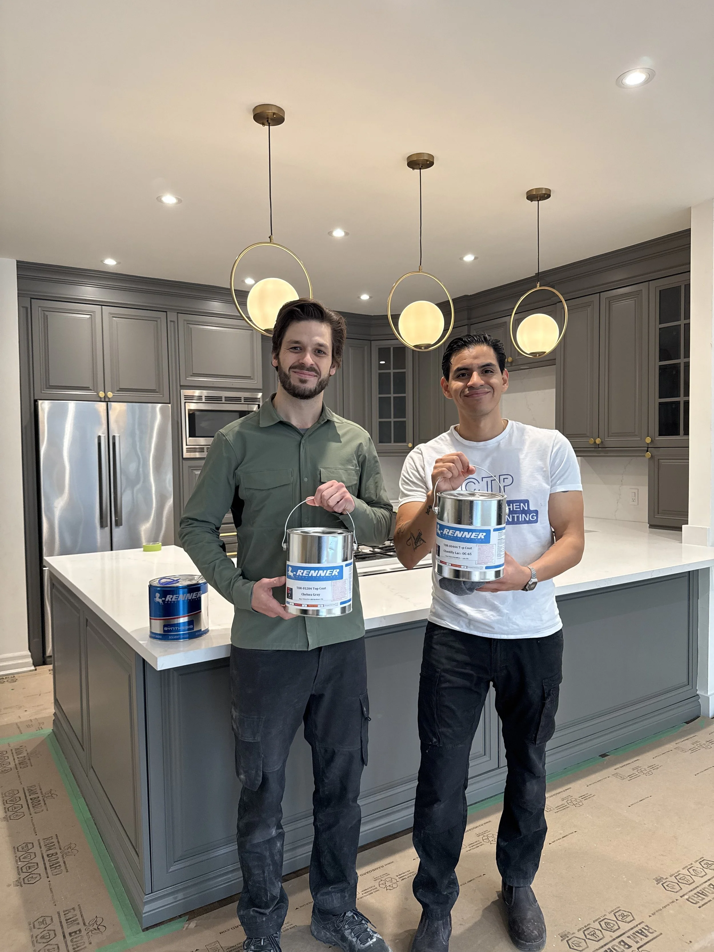 Two men standing in a modern kitchen, holding cans of paint. The kitchen has gray cabinets, a white countertop, and three hanging pendant lights.