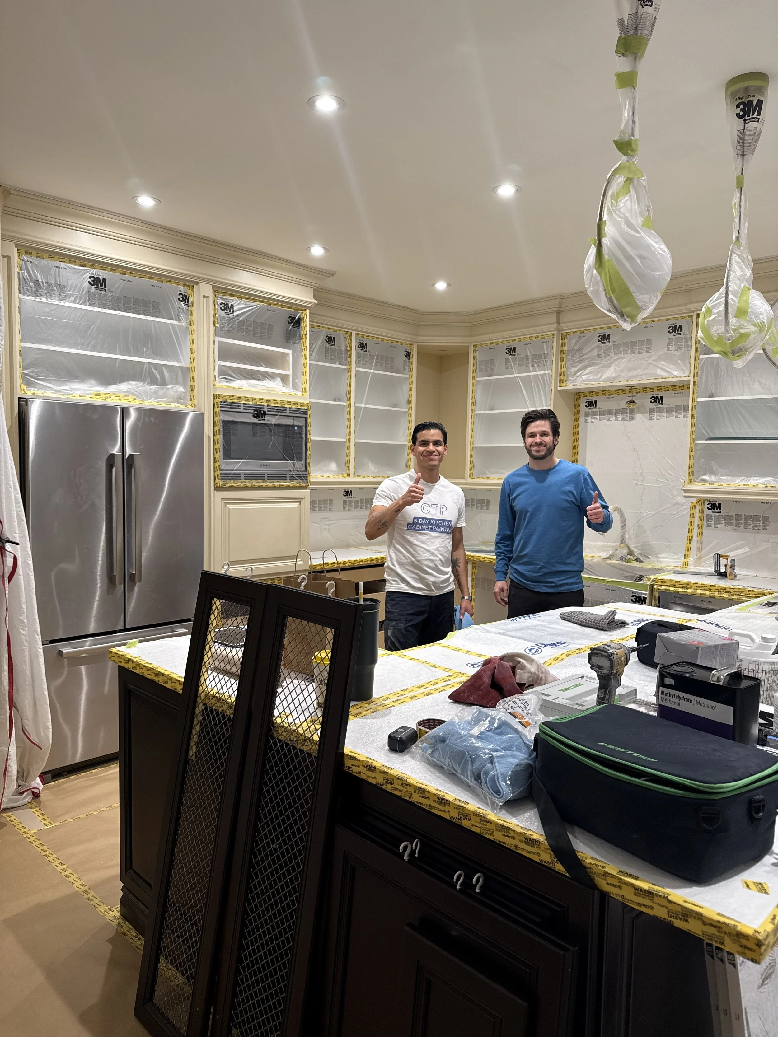 Two men standing in a kitchen under construction, smiling and giving thumbs-up. The kitchen cabinets are covered with protective plastic and tape, with construction tools and materials on the counter.