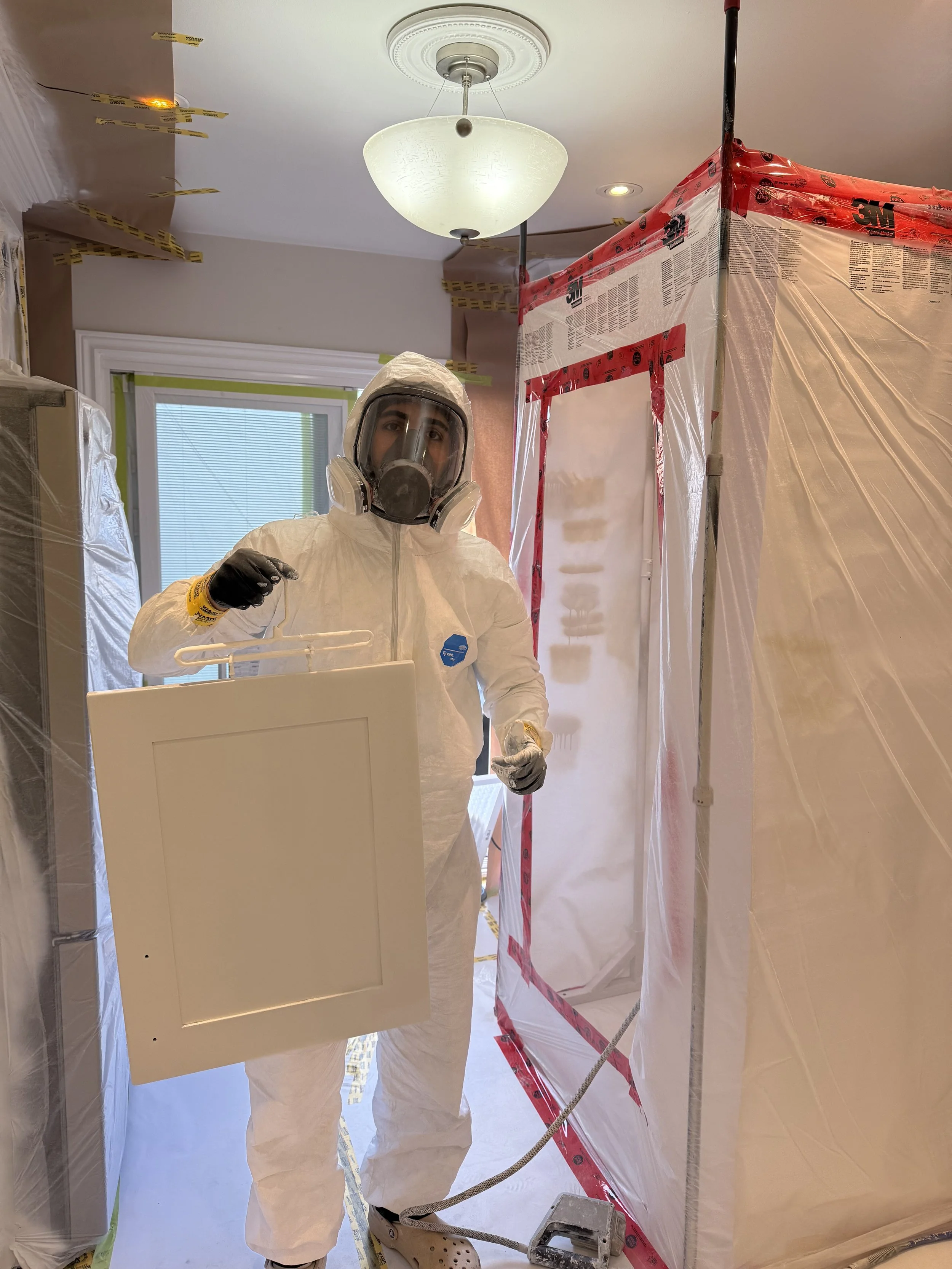 A person in full protective gear, including a hazmat suit, gloves, and a respiratory mask, standing inside a residential room with plastic sheeting and tape, holding a white panel, likely during a hazardous material or biohazard cleanup.