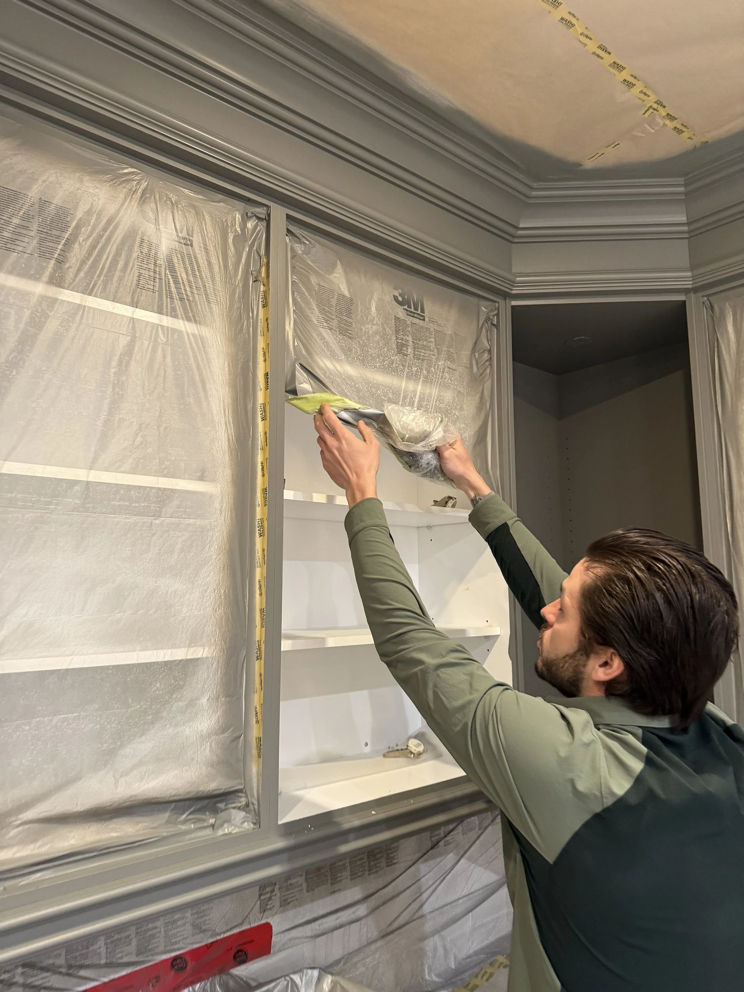 A man installing or repairing a kitchen cabinet, covering parts with protective plastic sheeting.