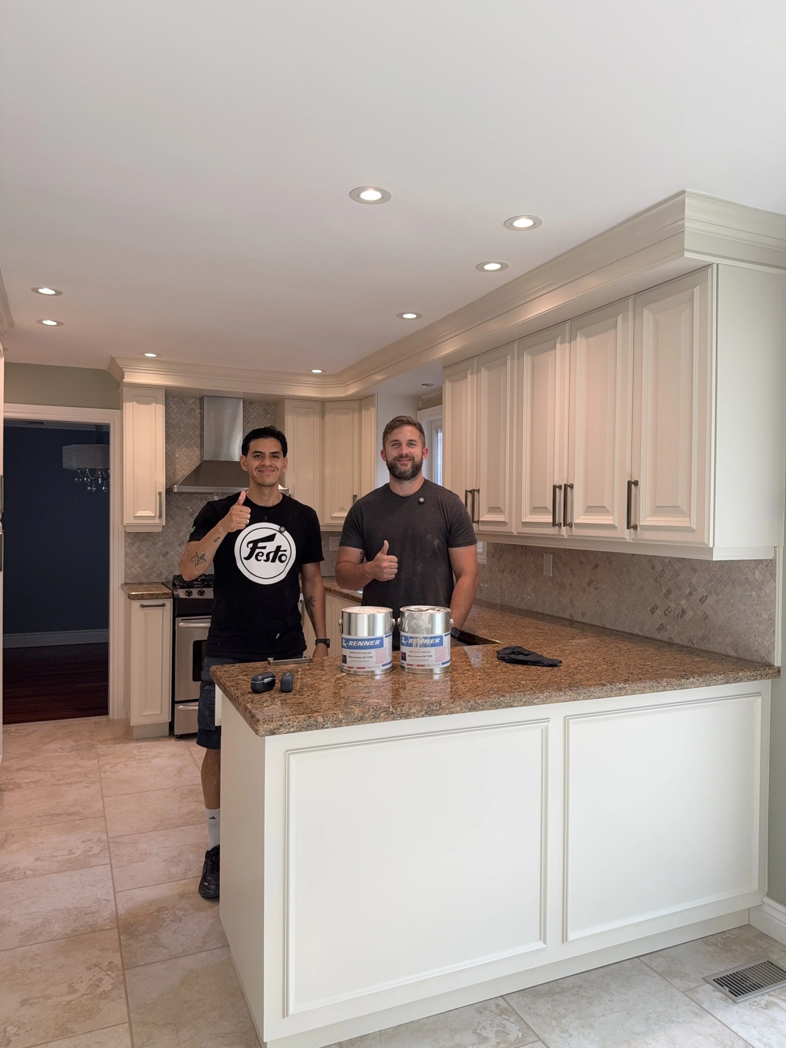 Two men standing in a kitchen behind a counter with paint cans, giving thumbs up.