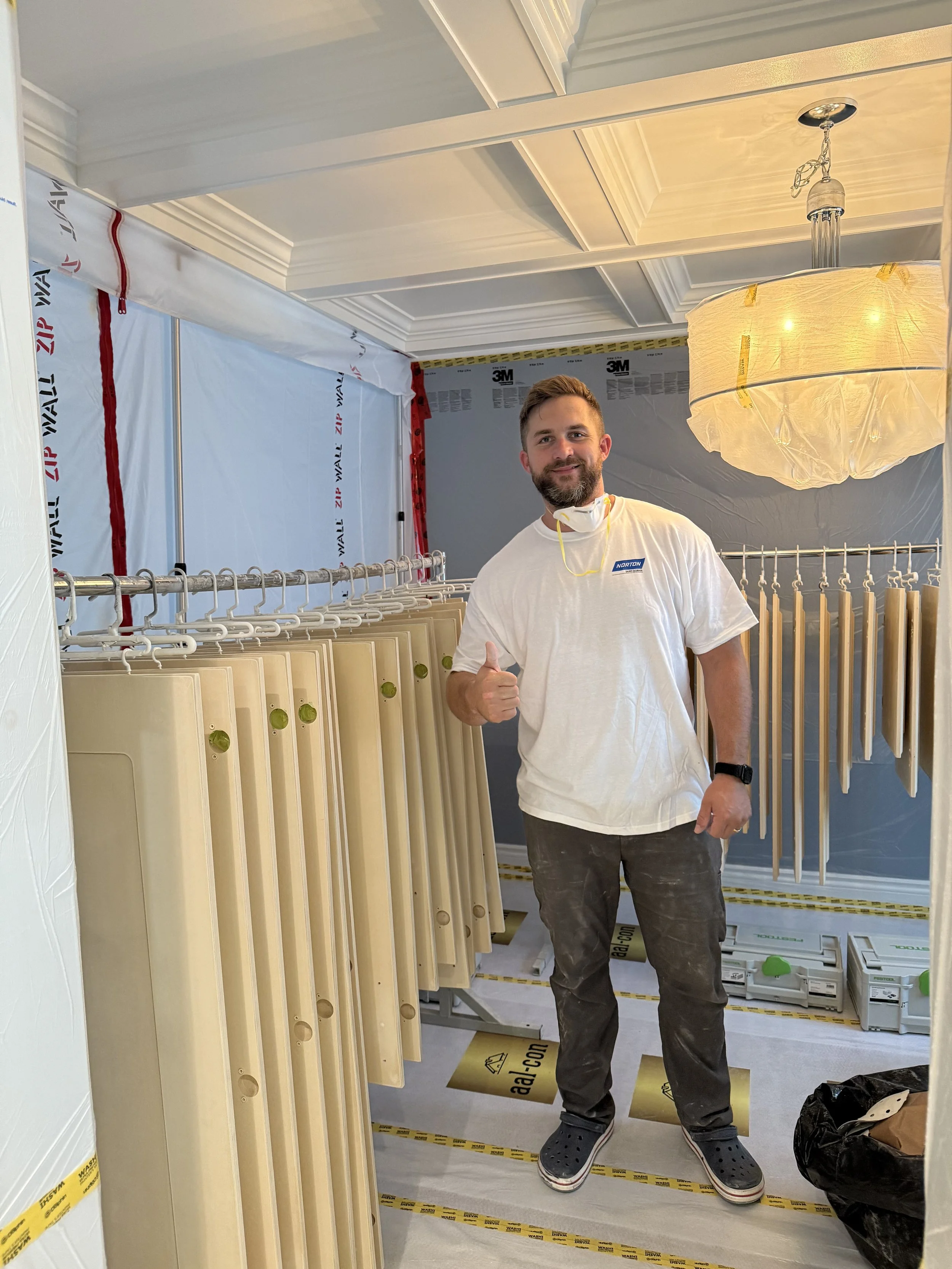 A man giving a thumbs-up in a room under construction, with drywall panels and insulation materials around him, and temporary protective coverings on the floor.