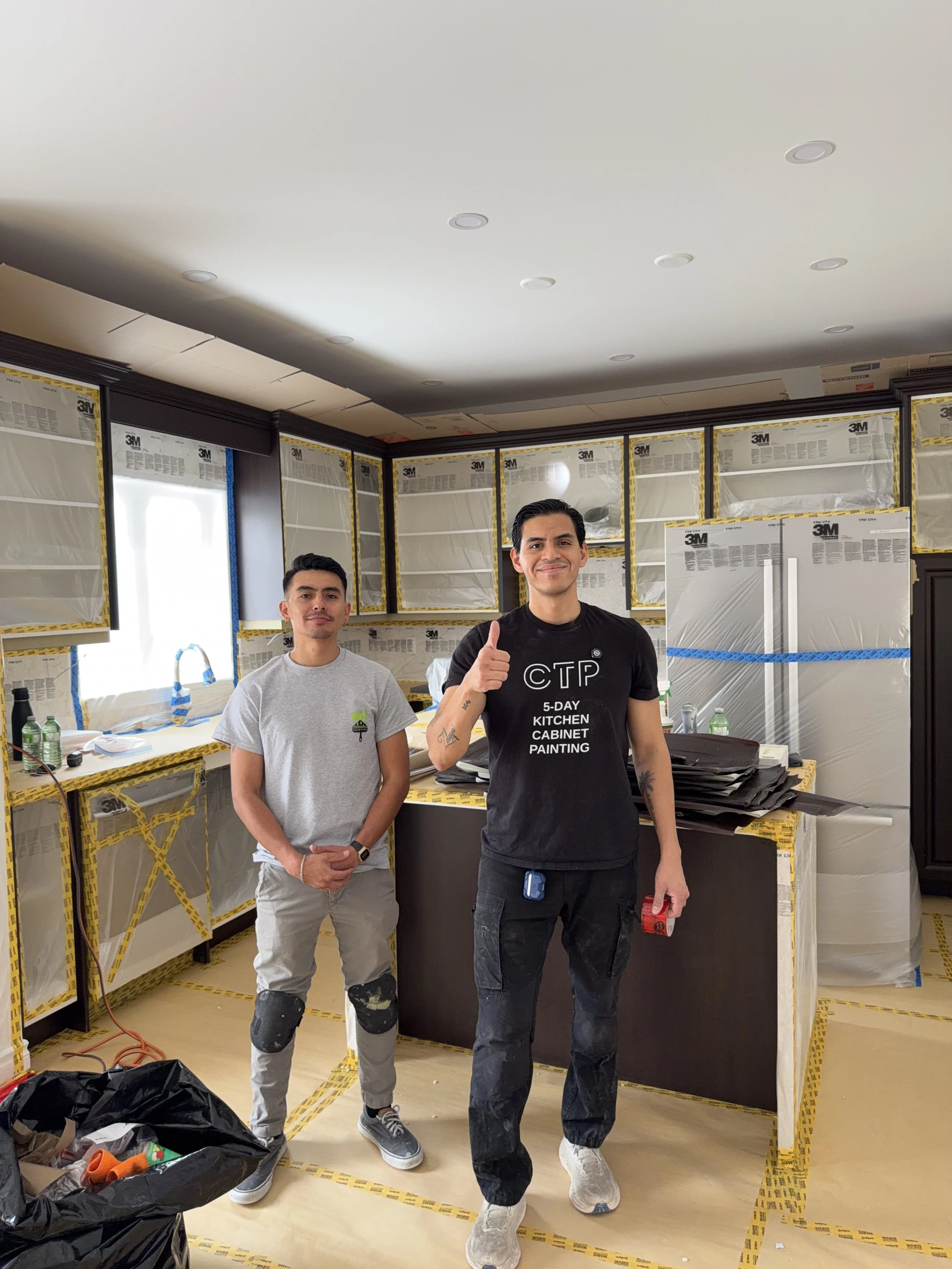Two men standing in a kitchen under renovation. The kitchen cabinets are covered with protective plastic and tape. One man is giving a thumbs-up and smiling, while the other stands with his hands clasped and looks at the camera.