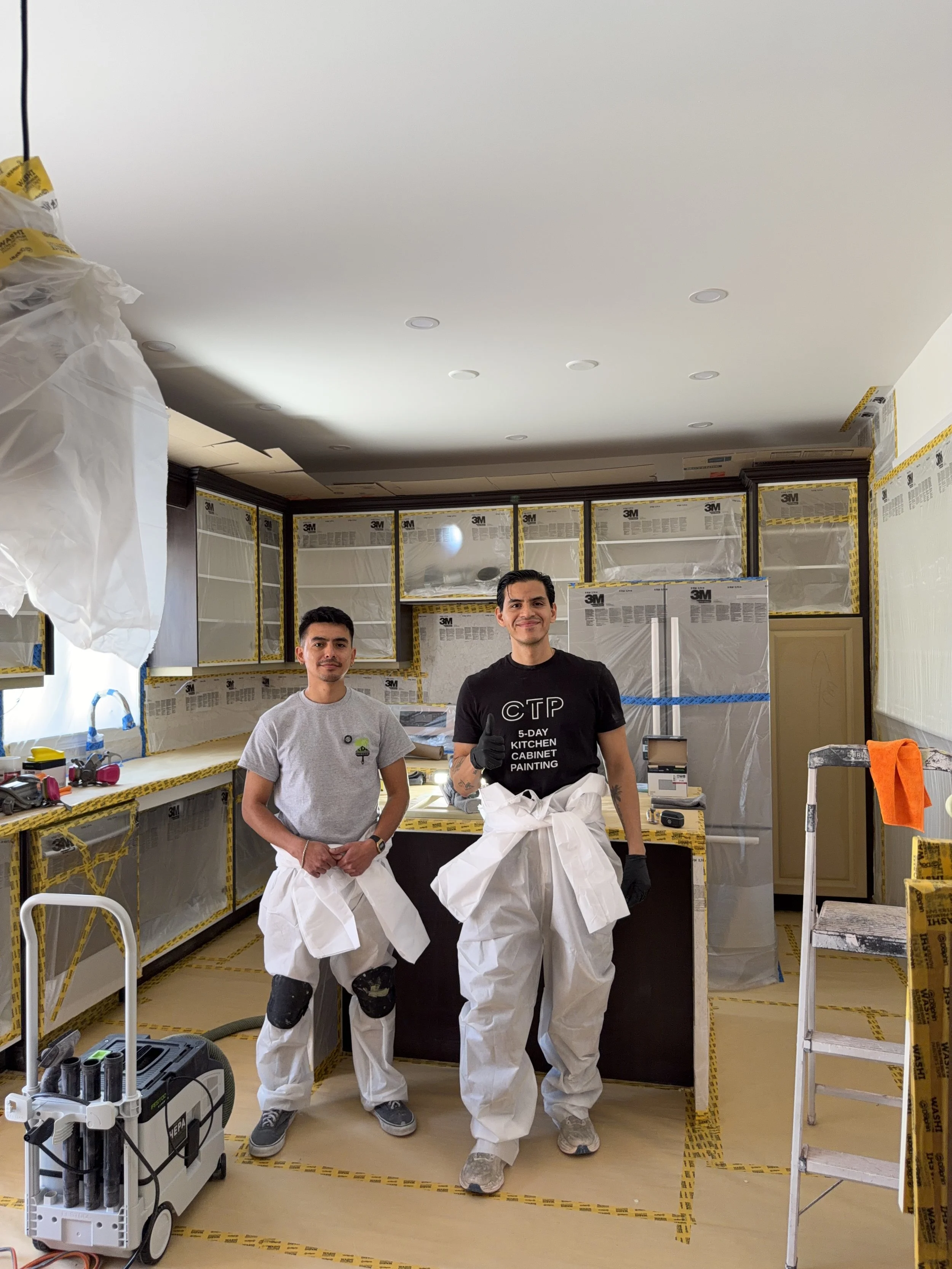 Two men in a kitchen under renovation, wearing white protective pants, with one smiling and giving a thumbs-up. The kitchen is covered with 3M painter's tape, and tools are visible around.