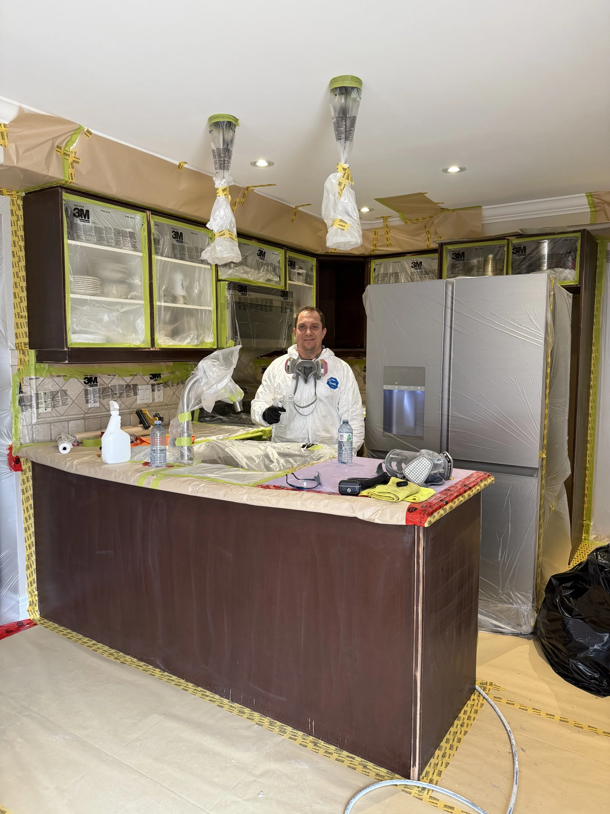 A man in protective work gear standing in a kitchen under renovation, smiling and giving a thumbs up. The kitchen is covered with painter's plastic and tape, with tools and water bottles on the counter.