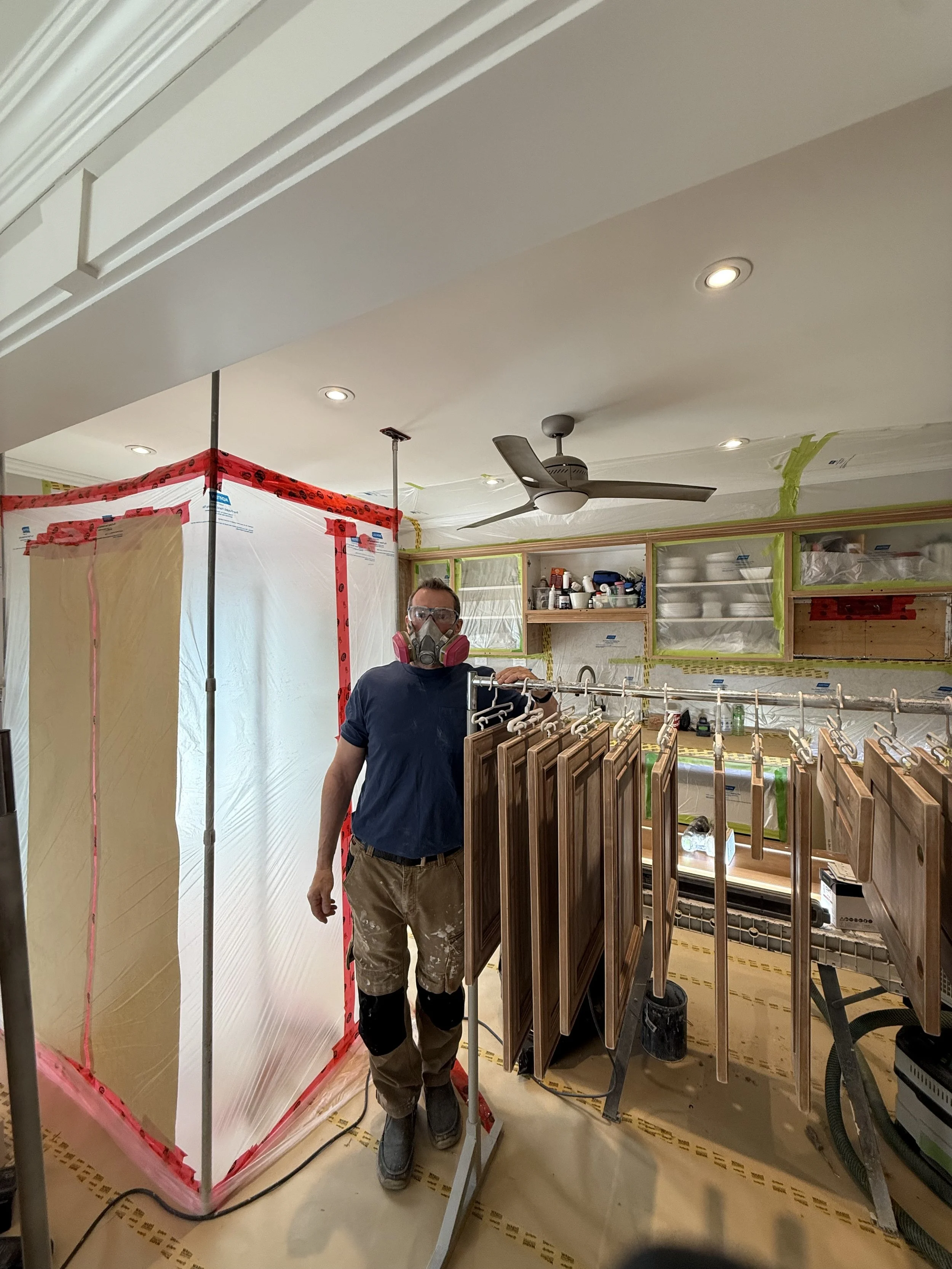 A person wearing a mask and safety glasses stands in a kitchen undergoing renovation. They are surrounded by hanging cabinet doors. The area is covered with plastic sheeting and tape, indicating ongoing construction work.