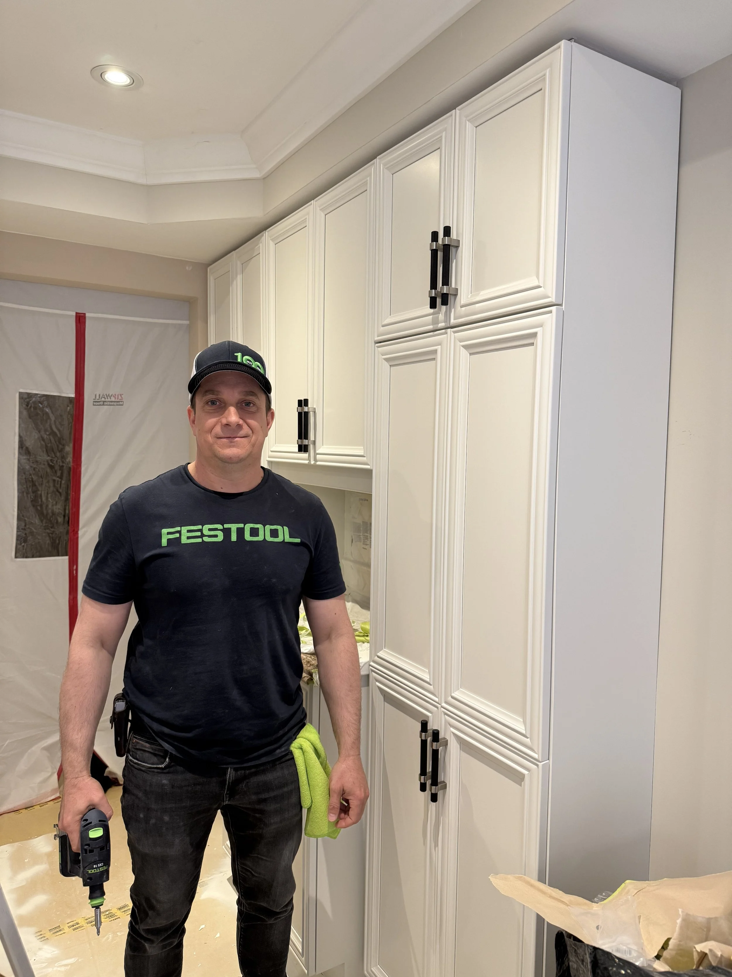 A man wearing a black FESTOOL T-shirt and cap, holding a power drill, standing in a kitchen with white cabinets and some construction materials around.
