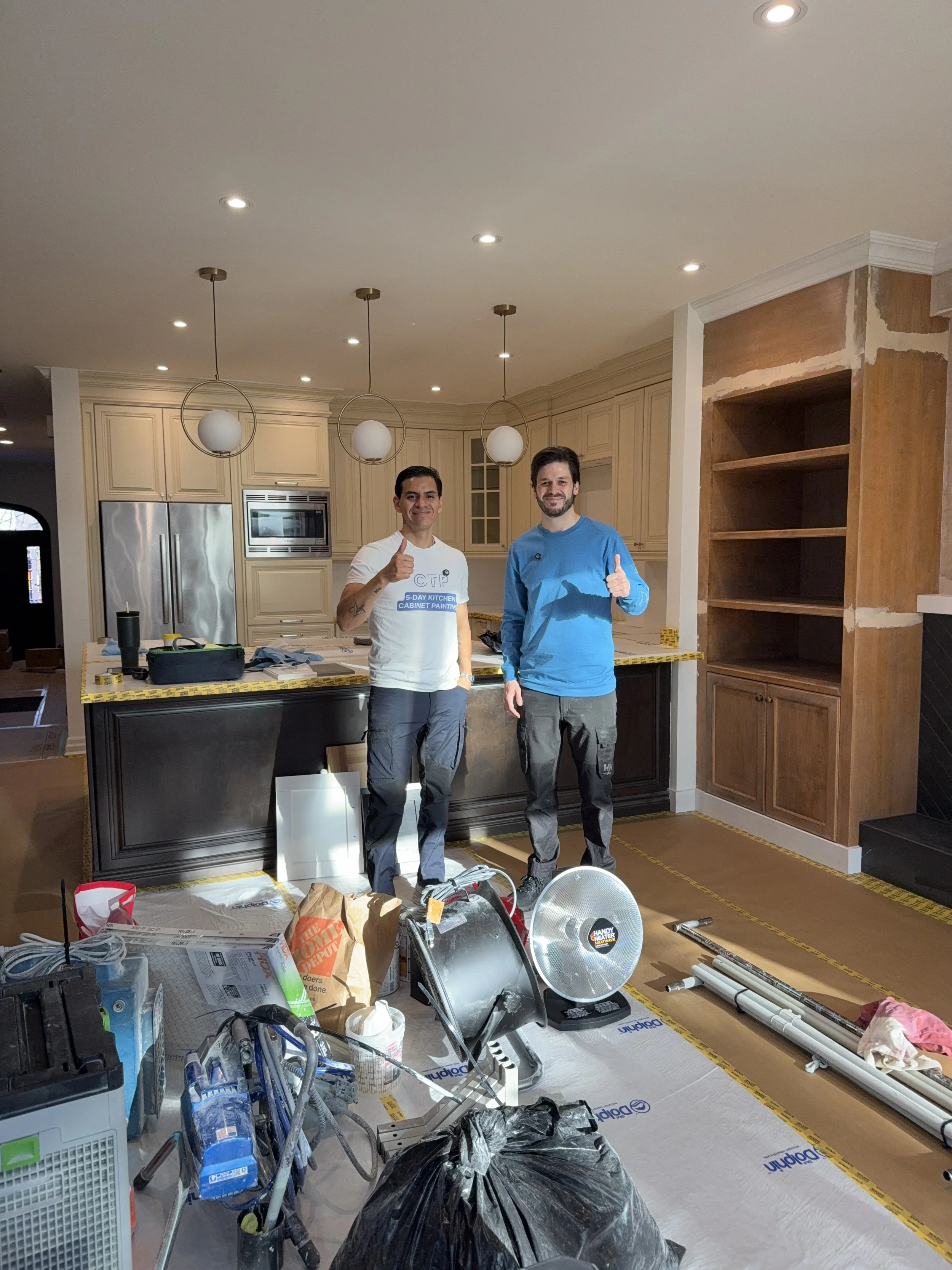 Two men standing inside a kitchen with ongoing renovation work, surrounded by tools and materials, giving thumbs up.