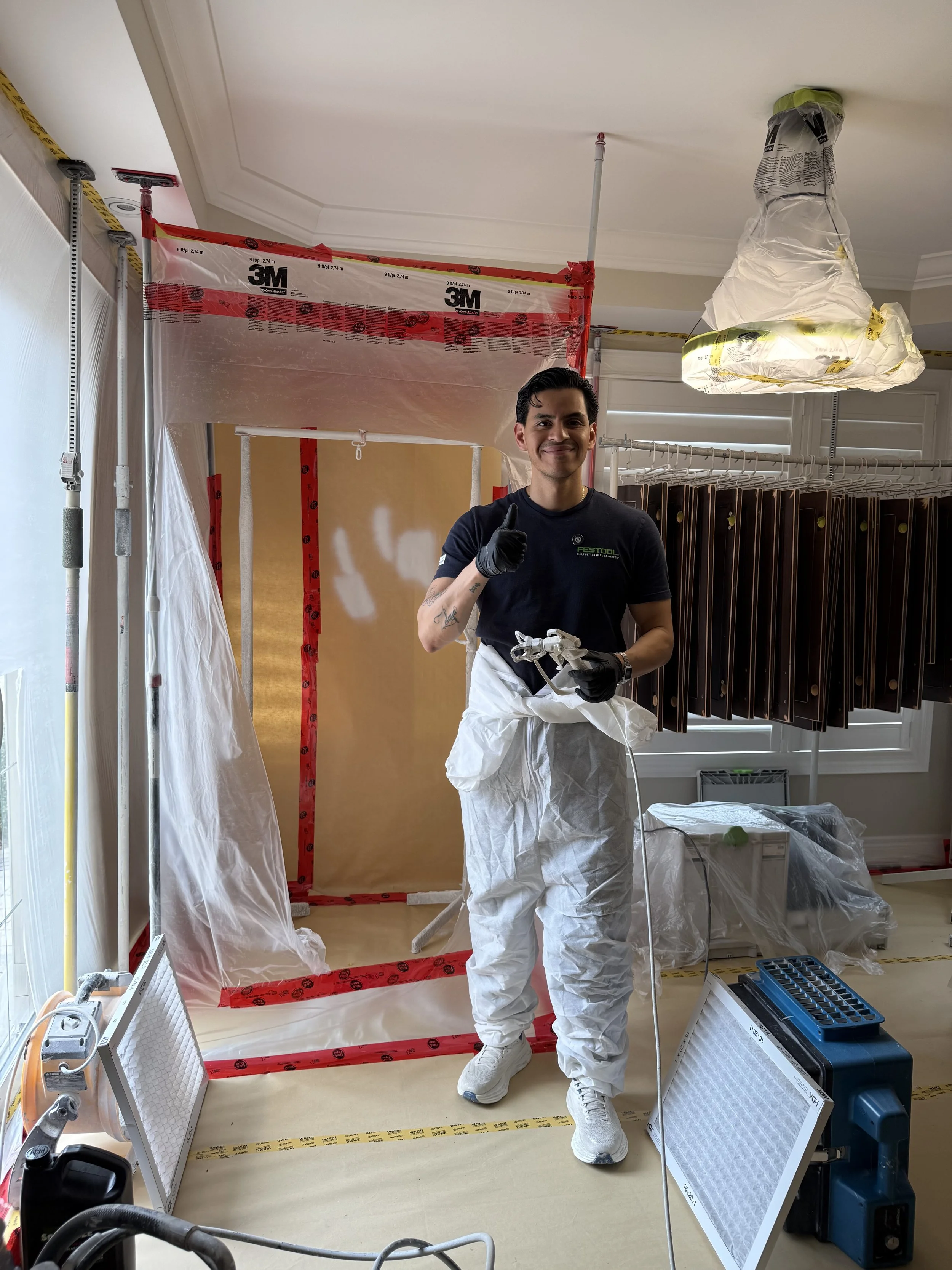 A man in protective gear holding a paint sprayer, standing in a room undergoing renovation with construction materials and equipment around him.