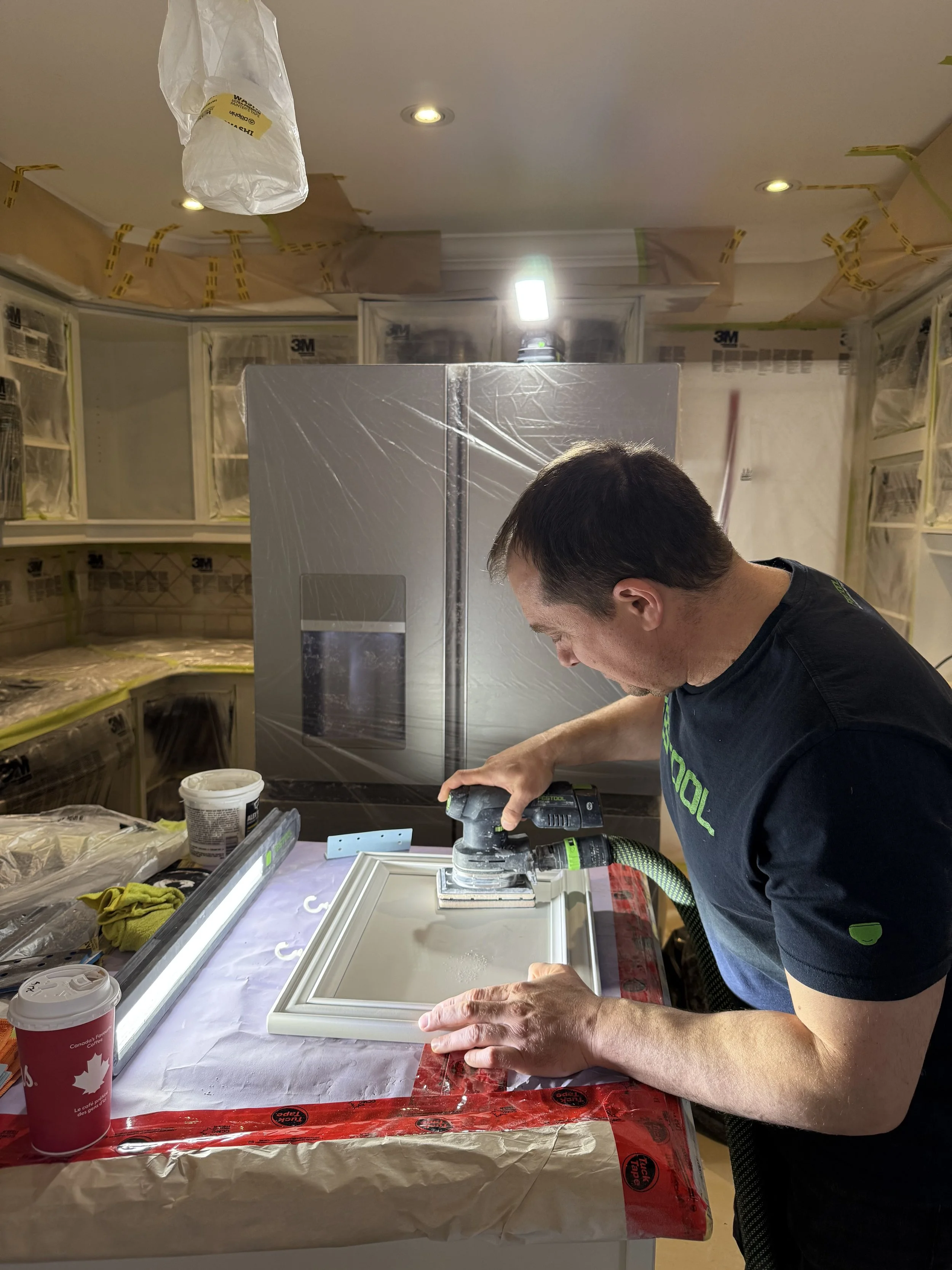 A man working on a cabinet door in a kitchen under renovation, using a handheld sander or polisher, with a large refrigerator covered in plastic in the background.