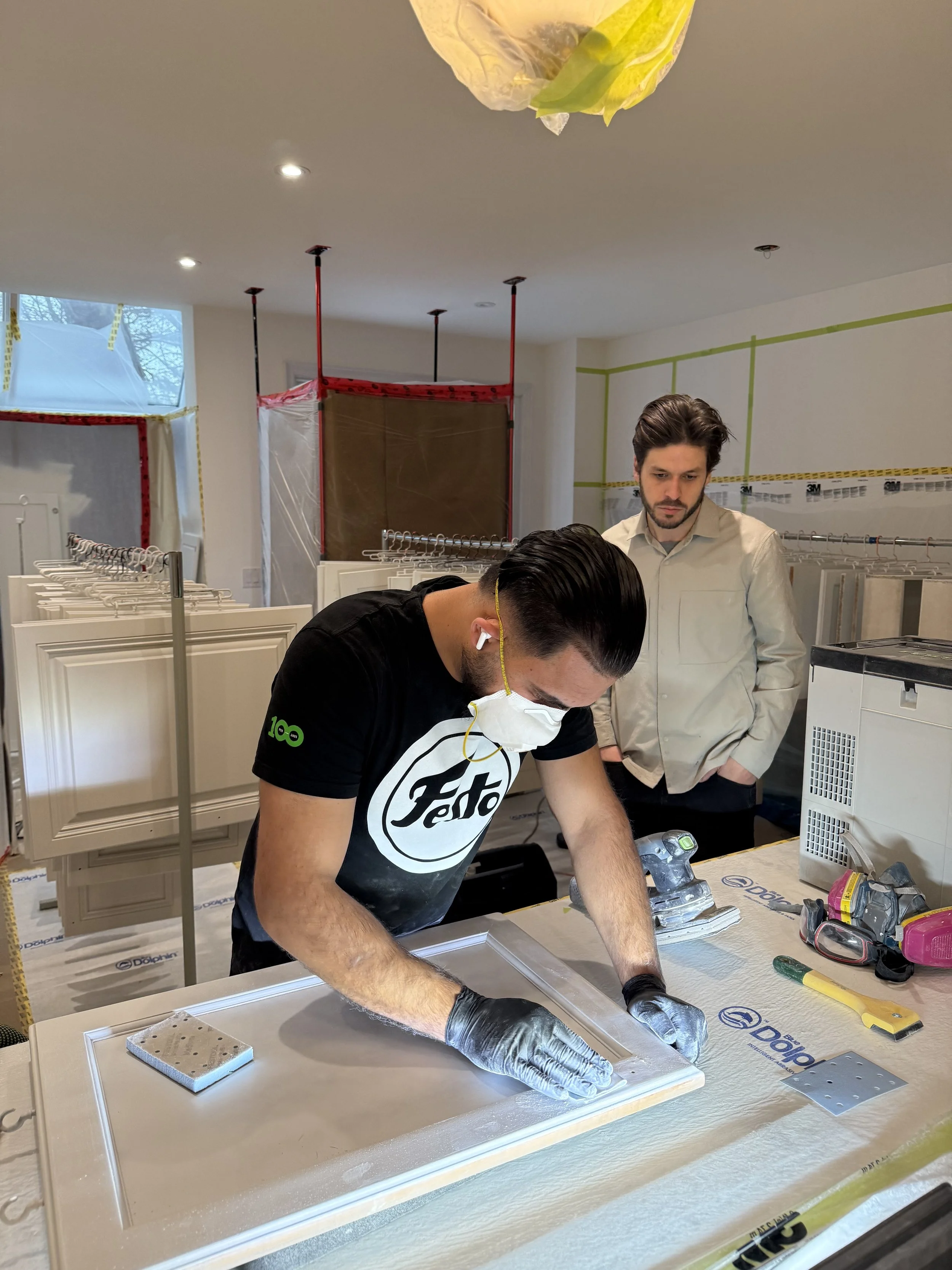 Two men working on a door during a renovation or construction project, one wearing a face mask, gloves, and wireless earbuds, and the other observing, in a room with construction materials and tools.