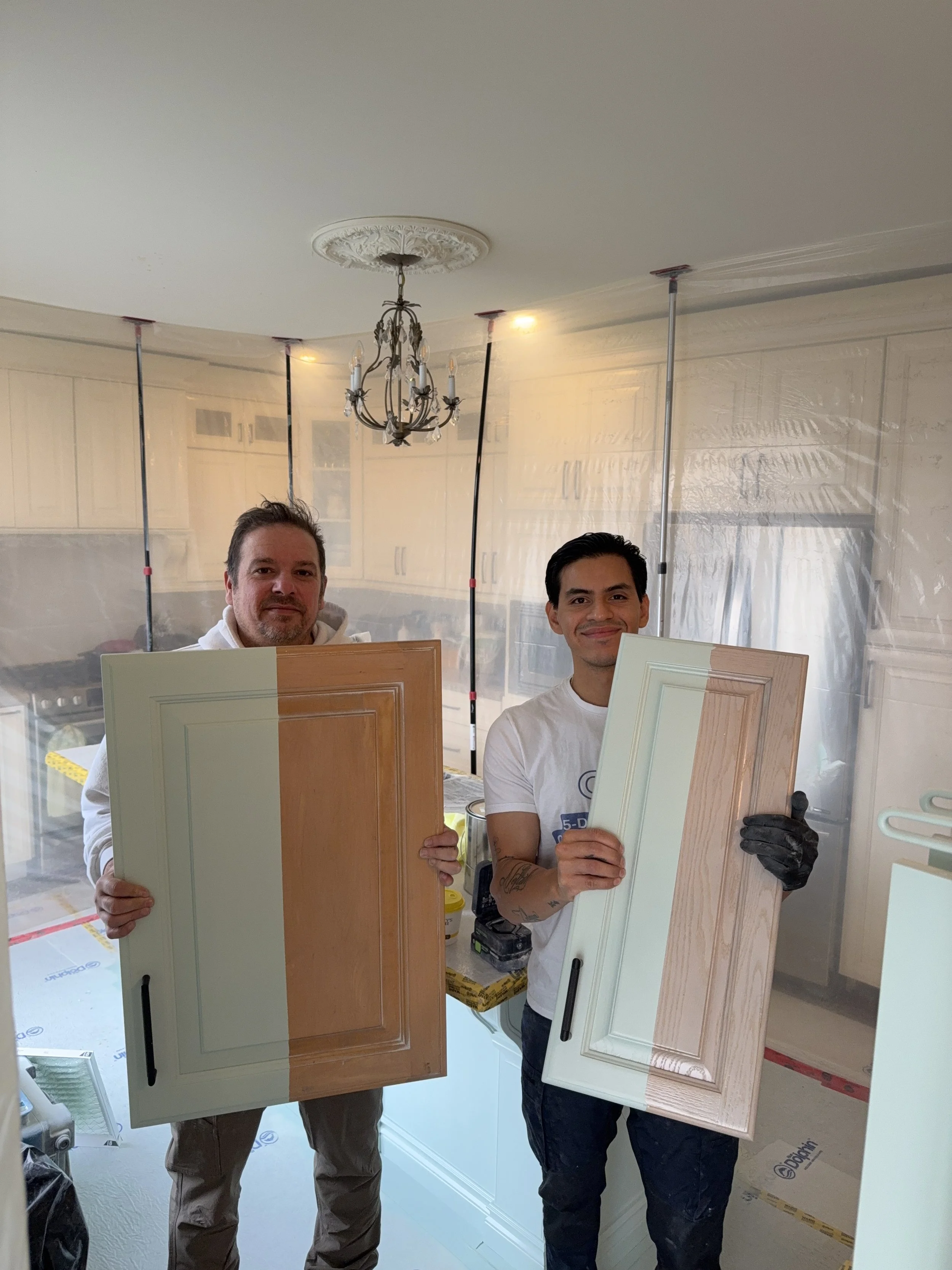 Two men are standing in a kitchen behind a plastic sheet, holding cabinet doors, one with light wood finish and the other painted white. They appear to be carpenters or contractors working on a renovation.