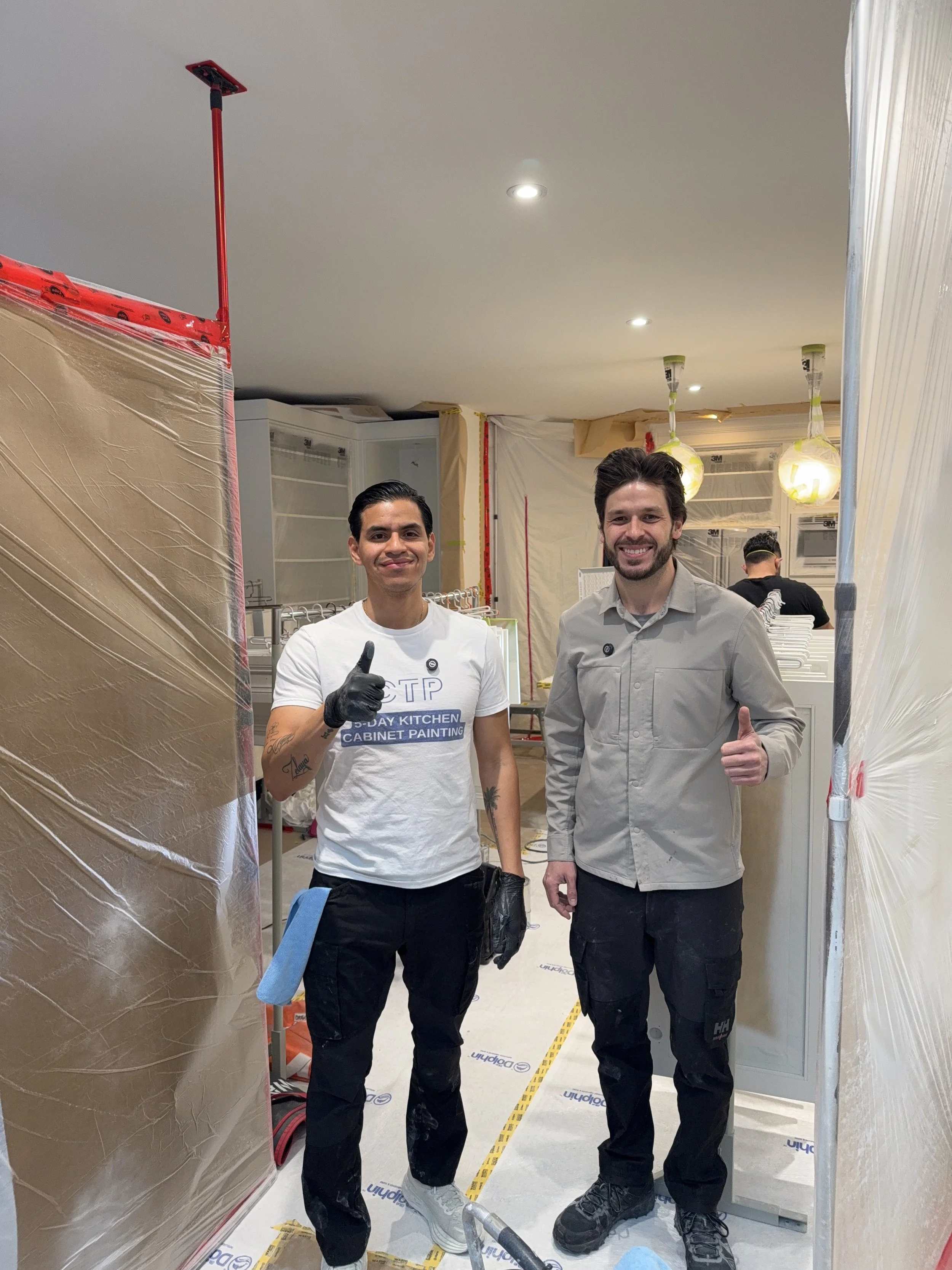 Two men smiling and giving thumbs up inside a home under renovation, with construction materials and plastic sheeting around.