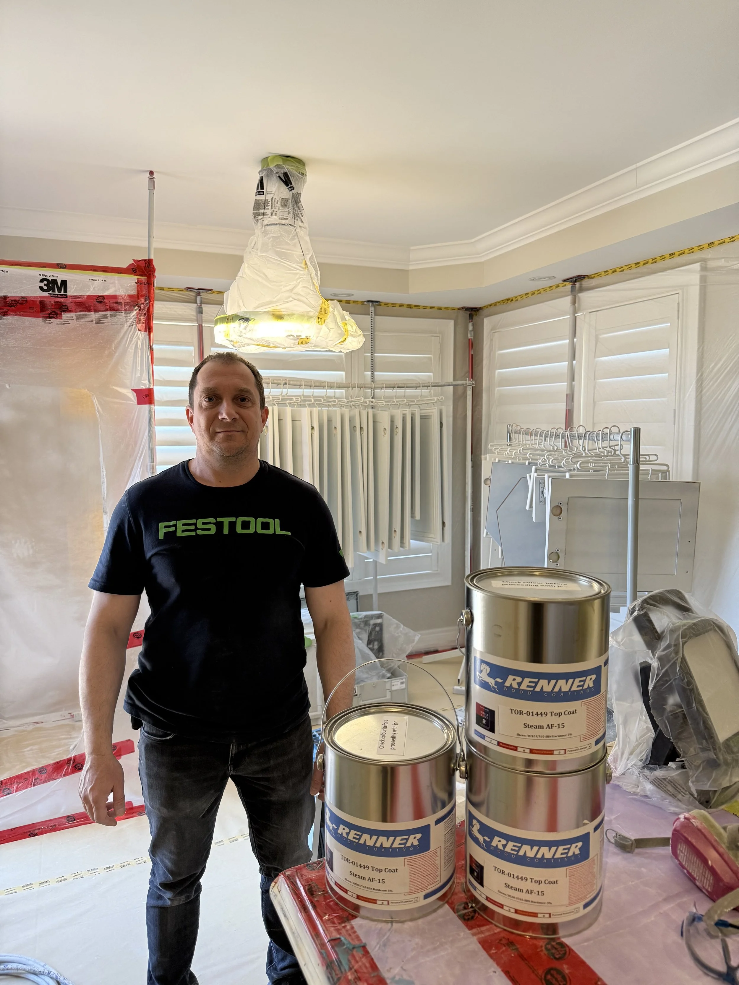 A man standing in a room that is under renovation, dressed in a black T-shirt with green text and dark jeans, surrounded by construction supplies including three cans of RENNER top coat paint, plastic sheeting, and window coverings.