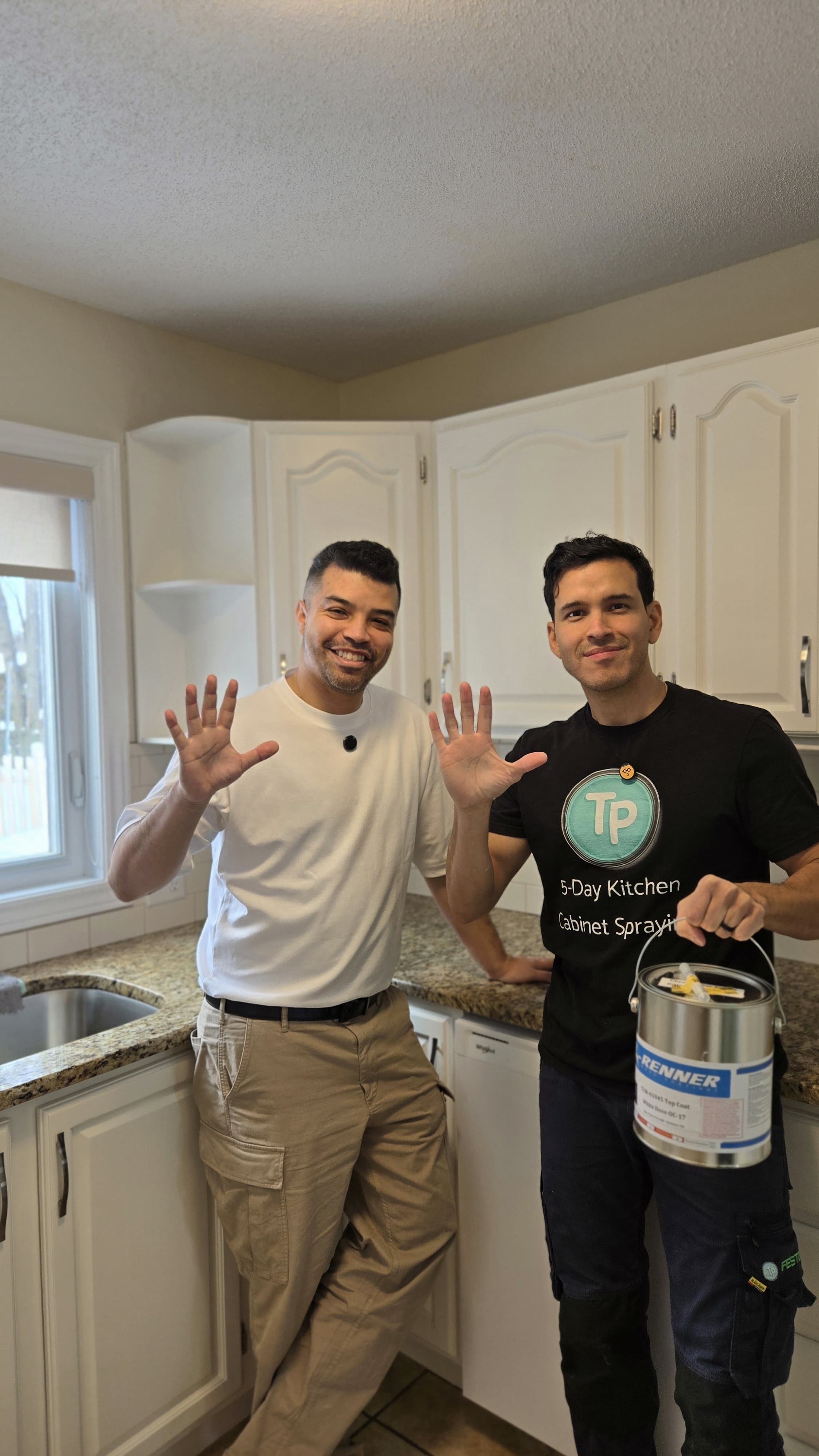 Two men in a kitchen smiling and waving at the camera, one holding a paint can, both casually dressed.