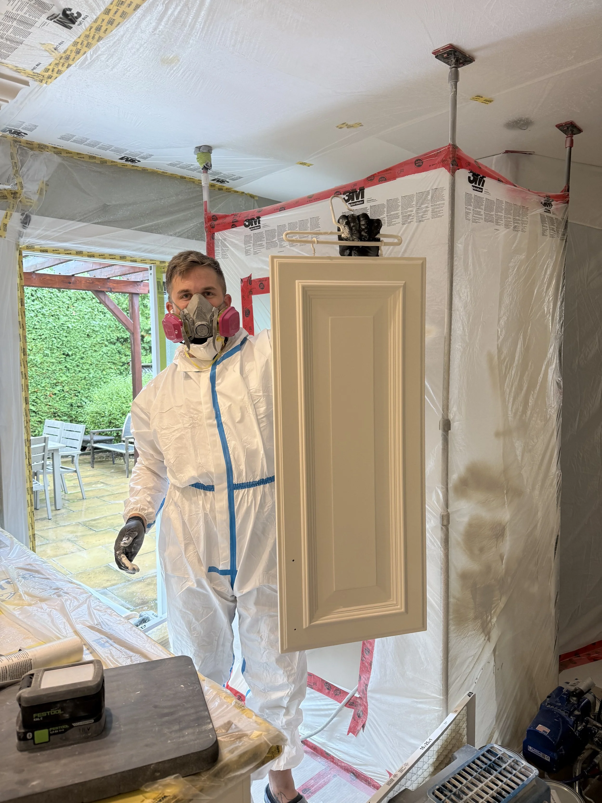 A man in protective gear holding a door panel inside a home renovation site with a backyard visible through an open door.