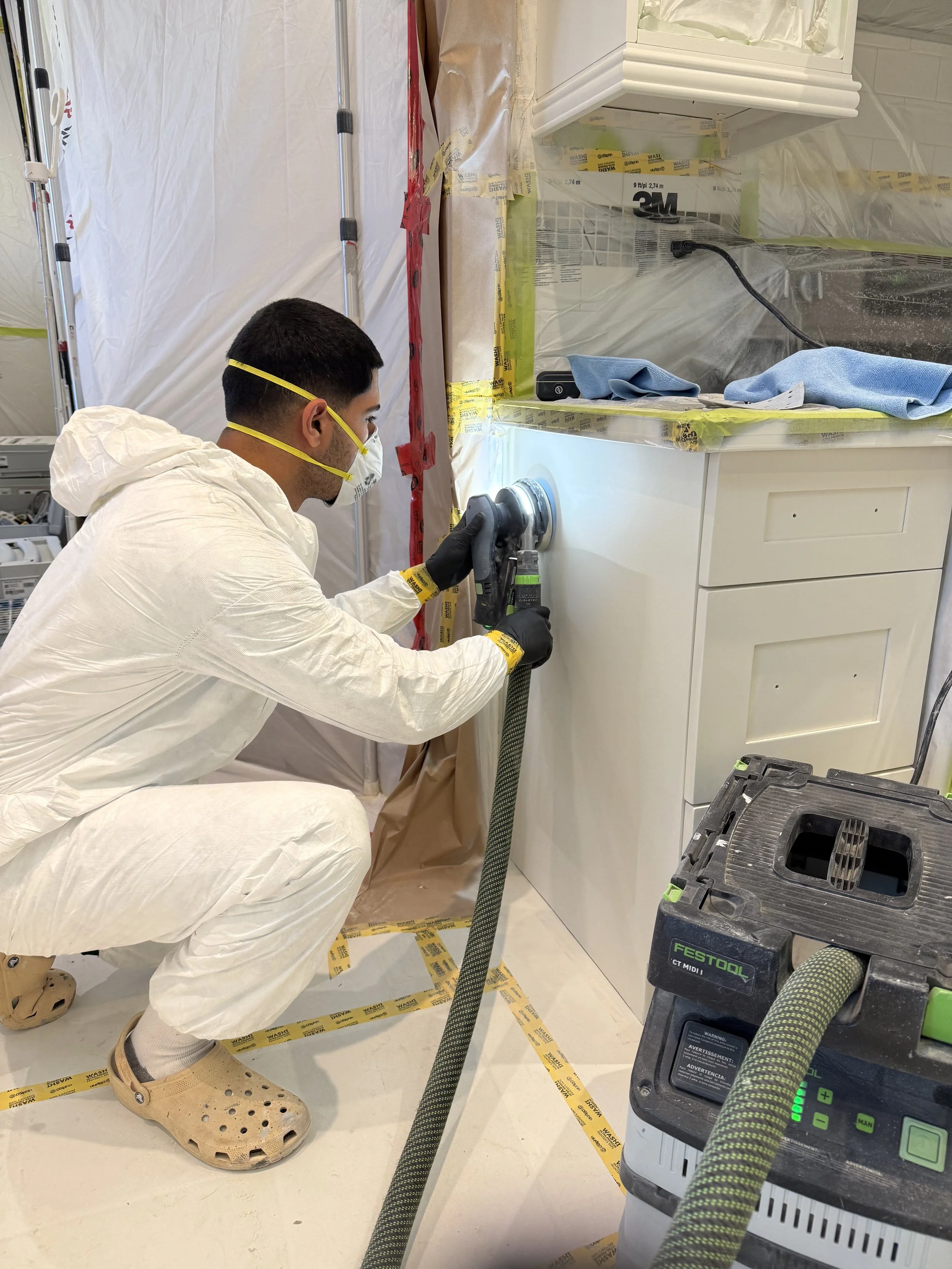 A worker wearing protective gear, including a mask, gloves, and a disposable suit, is sanding or polishing a white kitchen cabinet in a construction or renovation site. The workspace is surrounded by plastic sheeting and caution tape to protect the a