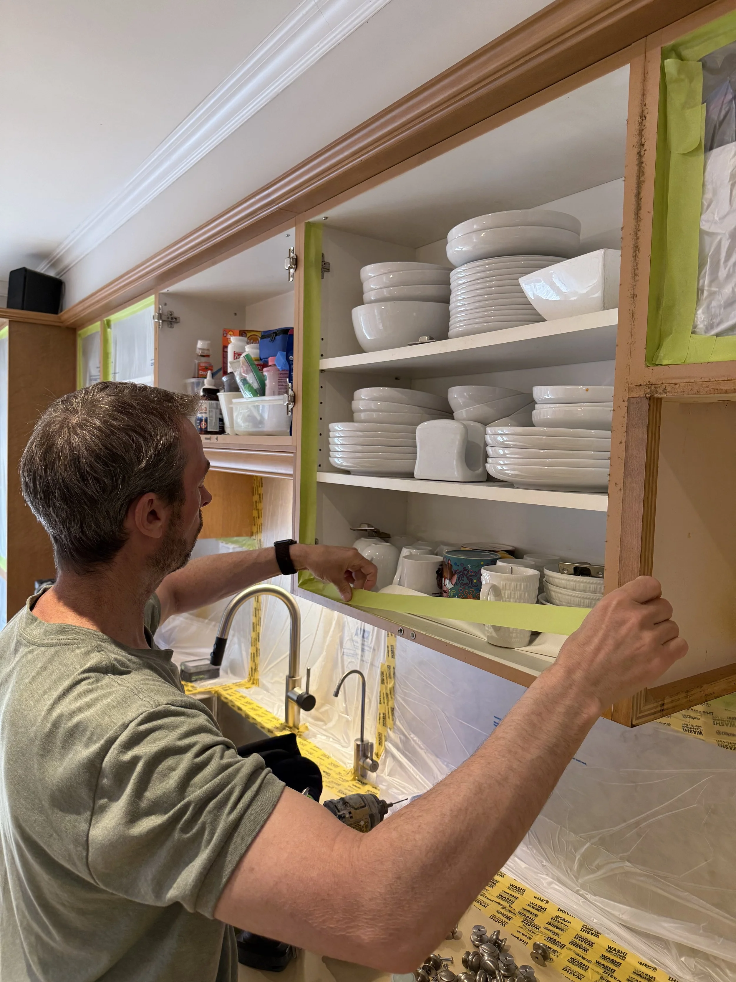 A man installing a kitchen cabinet above a sink, with open cabinet doors revealing stacked white dishes and kitchen supplies.