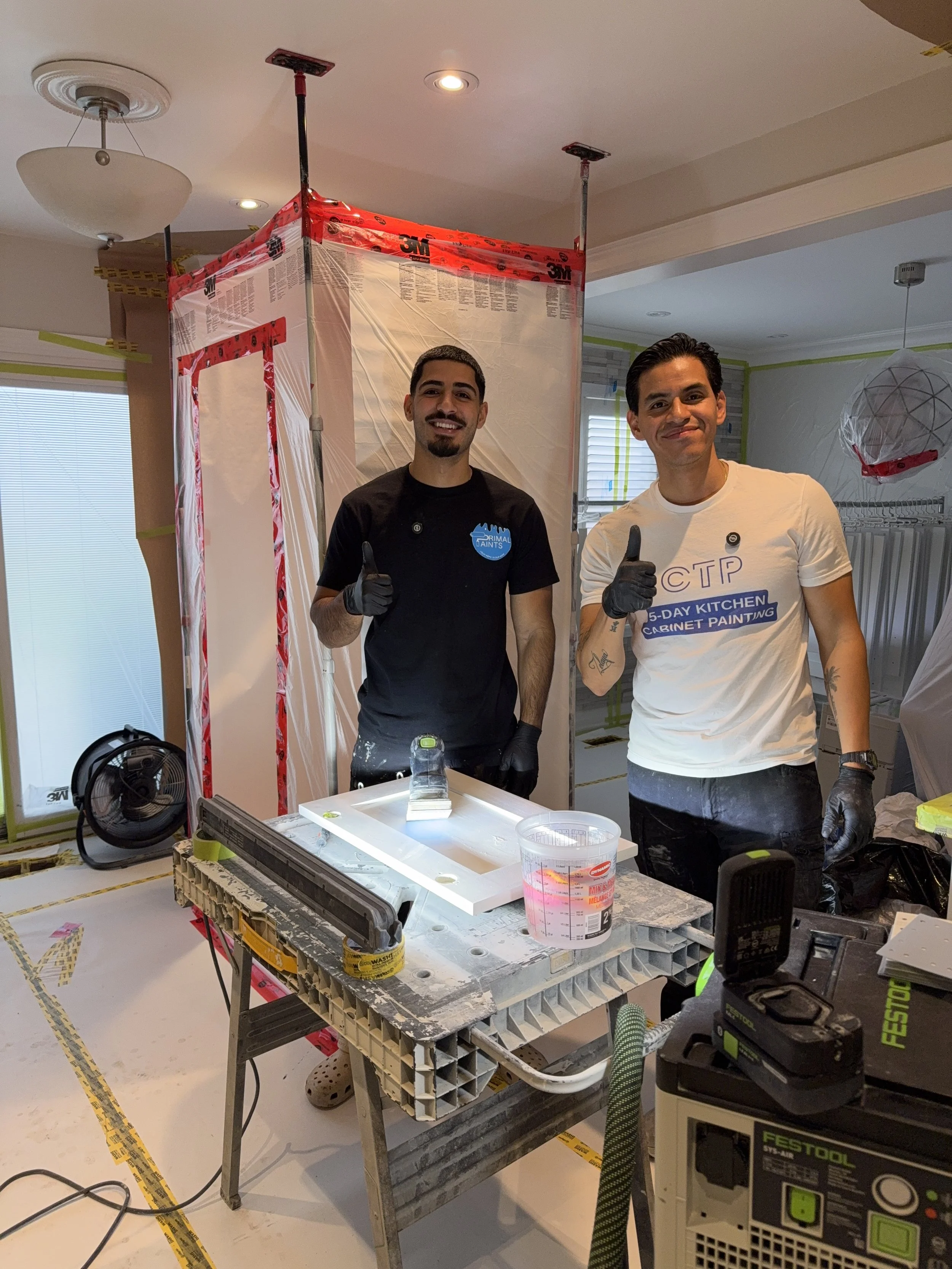 Two men wearing gloves and smiling give thumbs up in a room under renovation, with construction materials and tools on a table, and a plastic upgrade in progress.