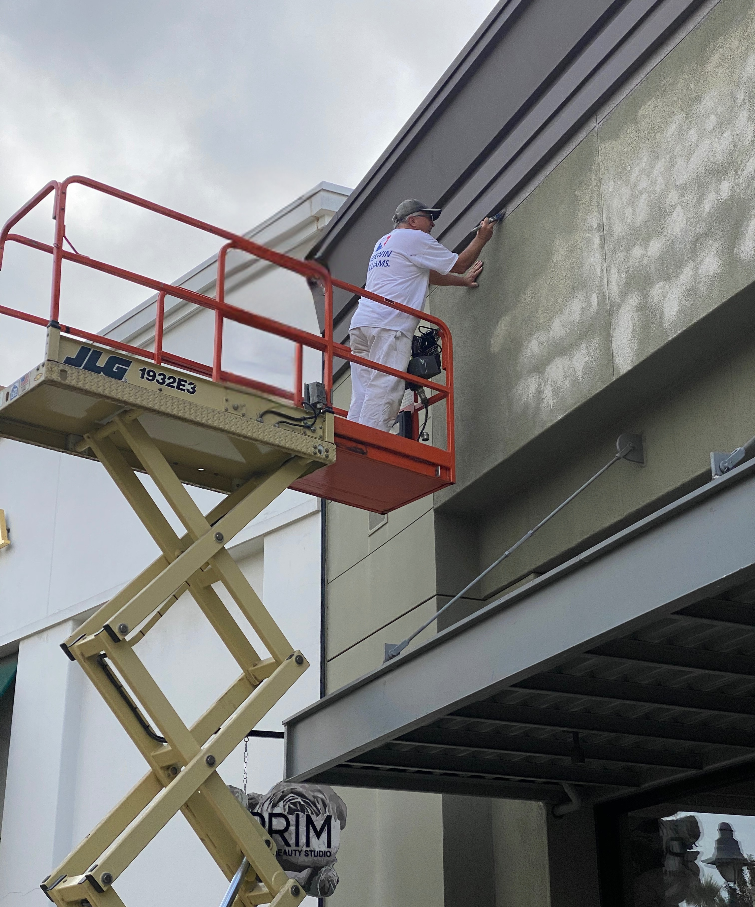 A worker stands on a cherry picker, painting the exterior wall of a building near the roofline.