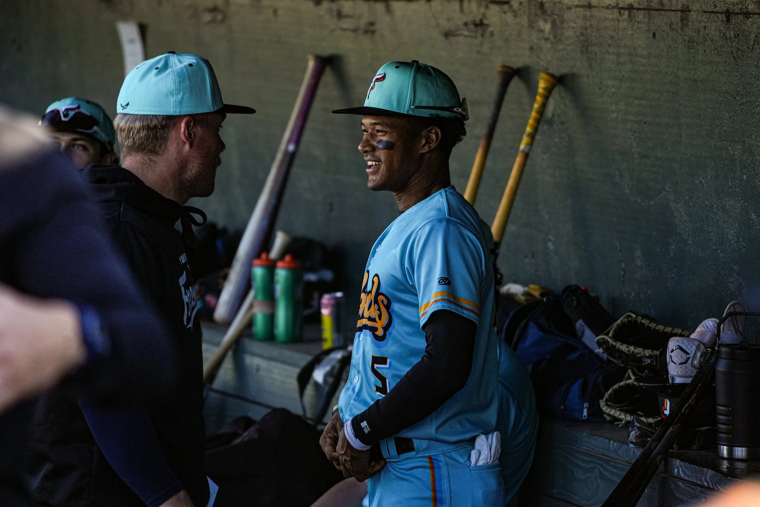 Two baseball players, one wearing a blue uniform and a cap, are smiling and talking in the dugout. Several baseball bats are leaning against the wall behind them.