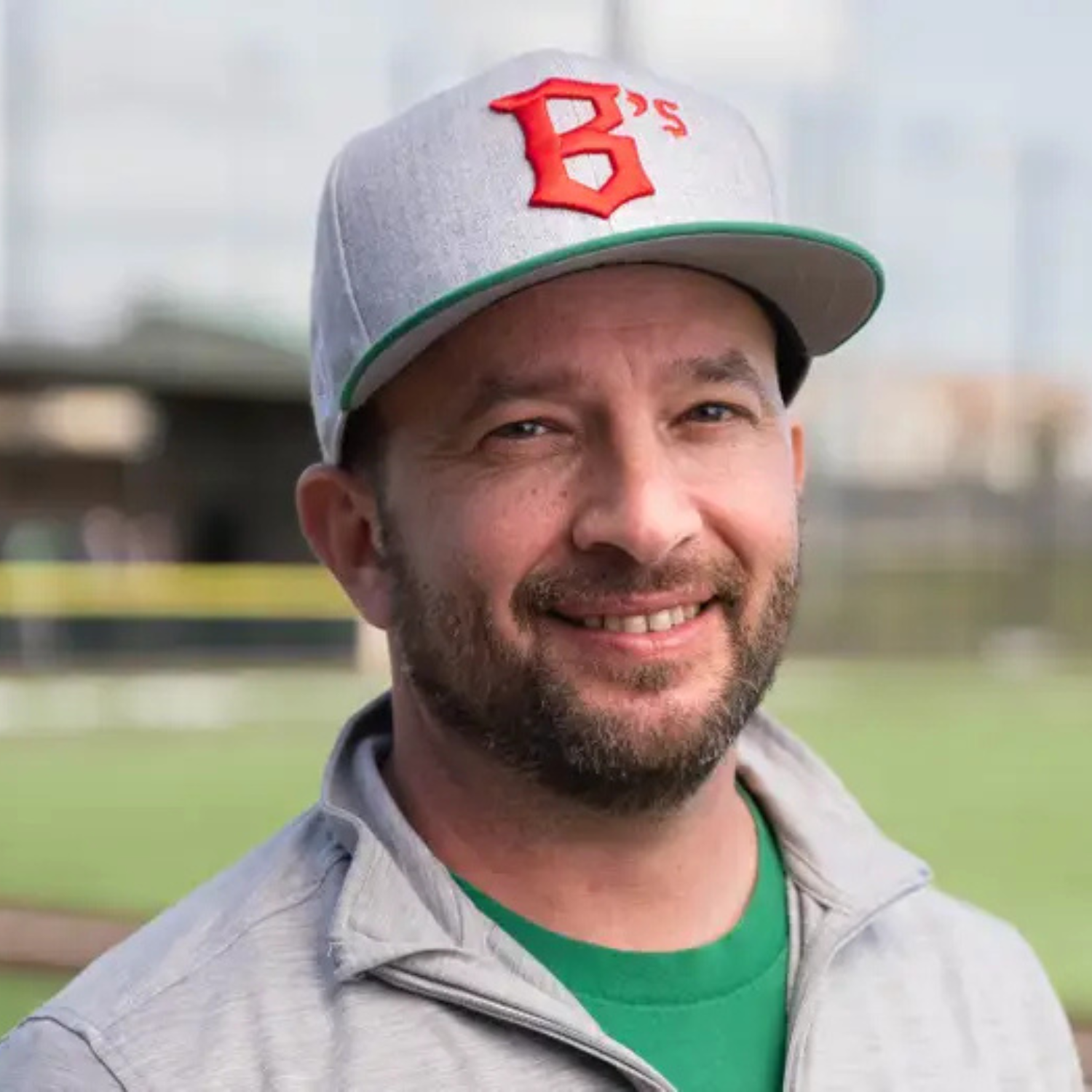Close-up of a smiling man wearing a baseball cap and athletic jacket, standing outdoors on a sports field.