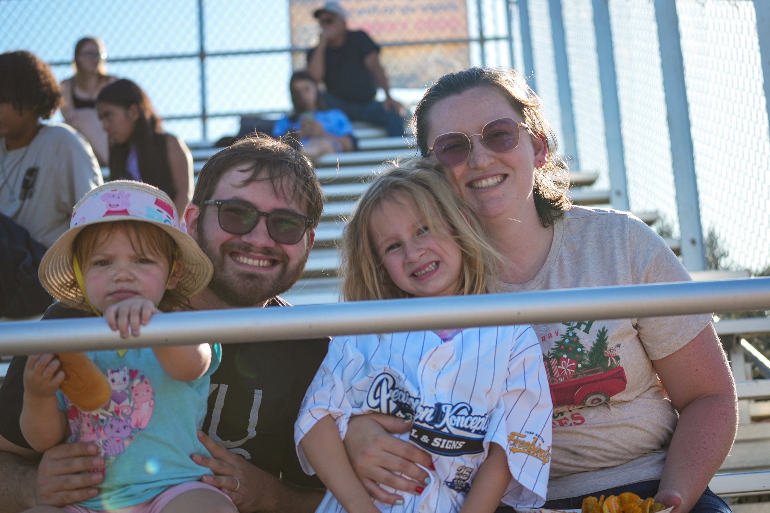 A joyful group of five people, including three children and two adults, smiling on stadium bleachers during daytime. The children are holding snacks and are dressed casually, with one wearing a sun hat. The background shows a chain-link fence and other spectators.