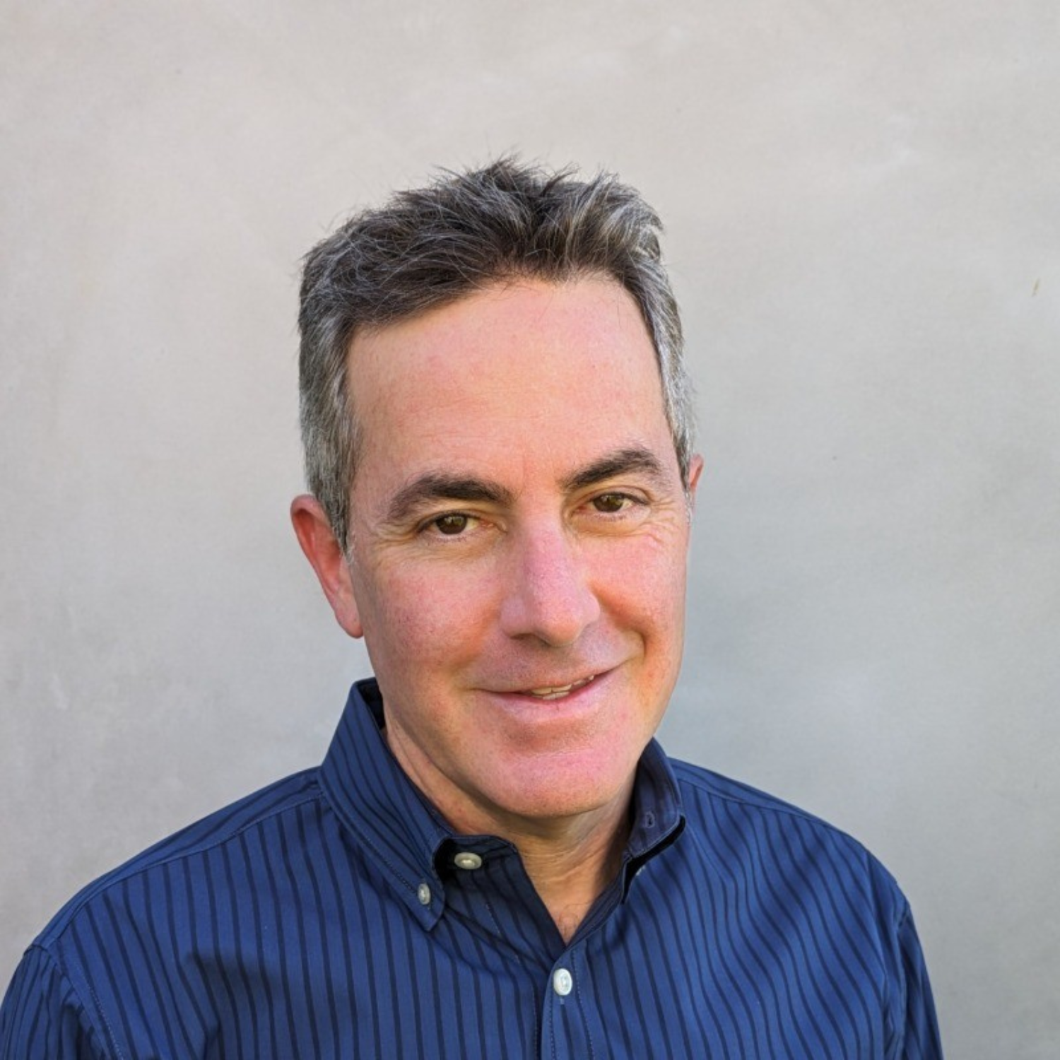 A man with gray hair and a blue striped shirt standing against a plain light-colored background.