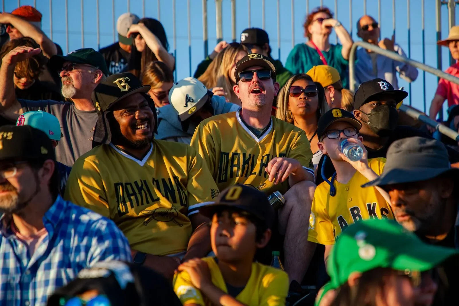 Crowd of baseball fans, some wearing Oakland Ballers jerseys and hats, enjoying a game outdoors on a sunny day.