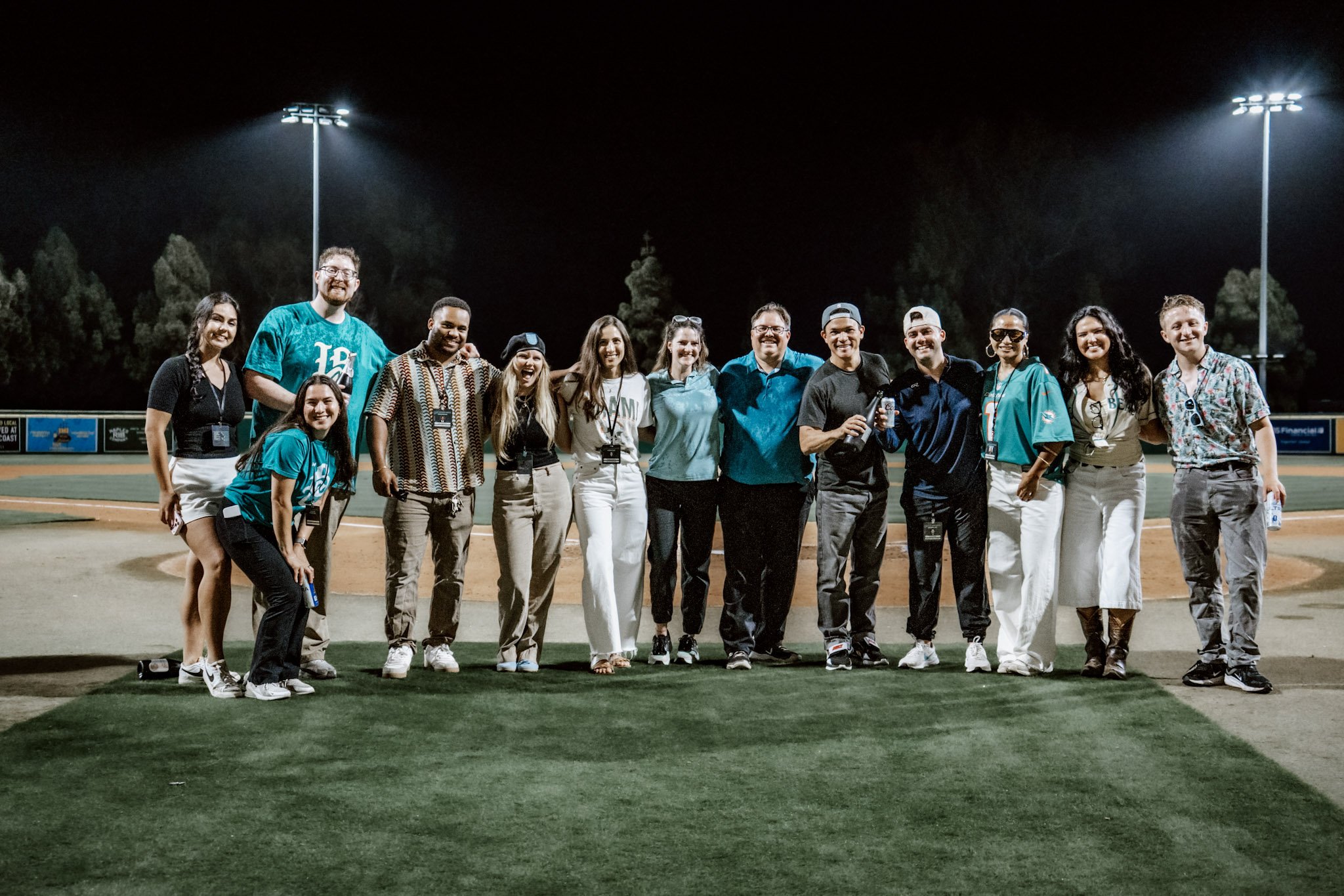 Group of people standing on a baseball field at night, smiling and posing for a photo. There are floodlights illuminating the field and trees in the background.
