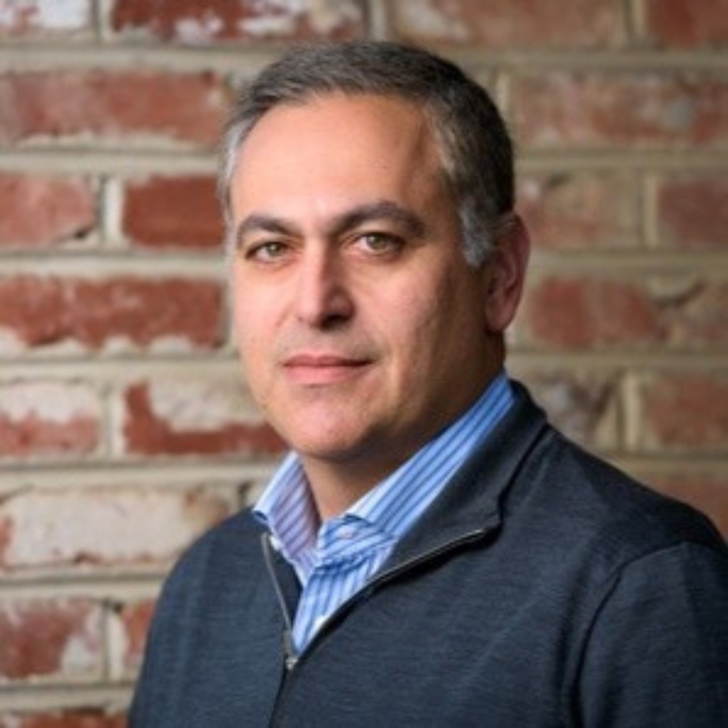 A man with grayish hair and a slight beard, wearing a blue and white striped shirt and a gray zip-up sweater, standing in front of a brick wall.