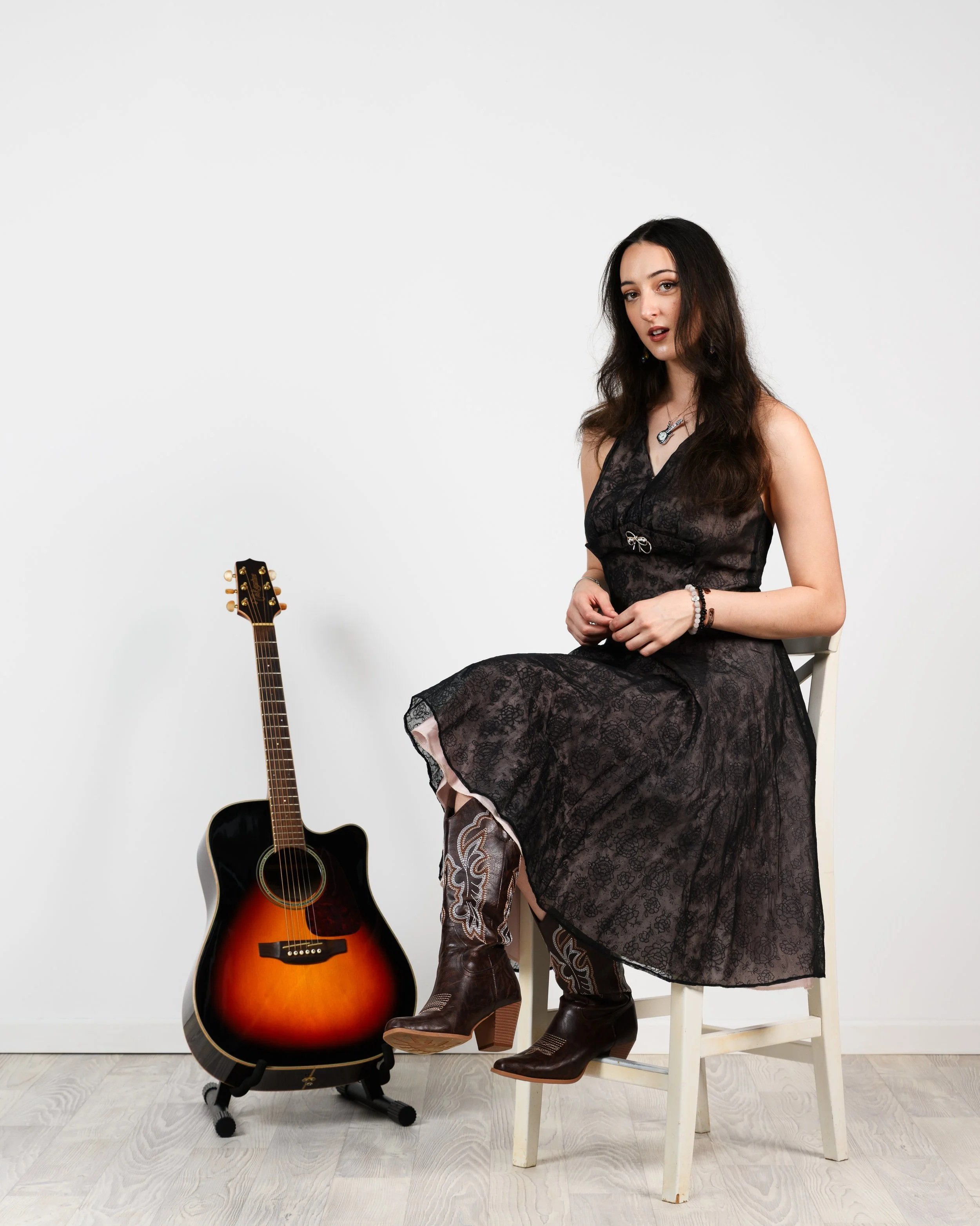 Shanleigh Rose with long dark hair wearing a black lace dress and cowboy boots sitting on a white chair next to a sunburst acoustic guitar on a stand against a plain white background.