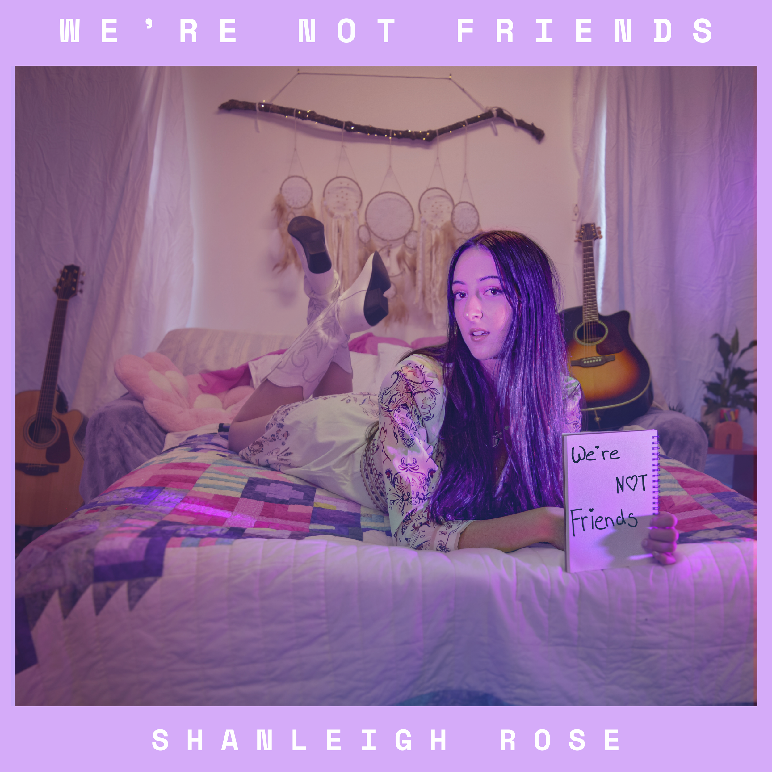 "We're Not friends" Cover art. Shanleigh Rose lying on her stomach on a bed, holding a notebook that says "We're NOT Friends." She is in a decorated bedroom with guitars on the wall and dreamcatchers hanging behind her. 